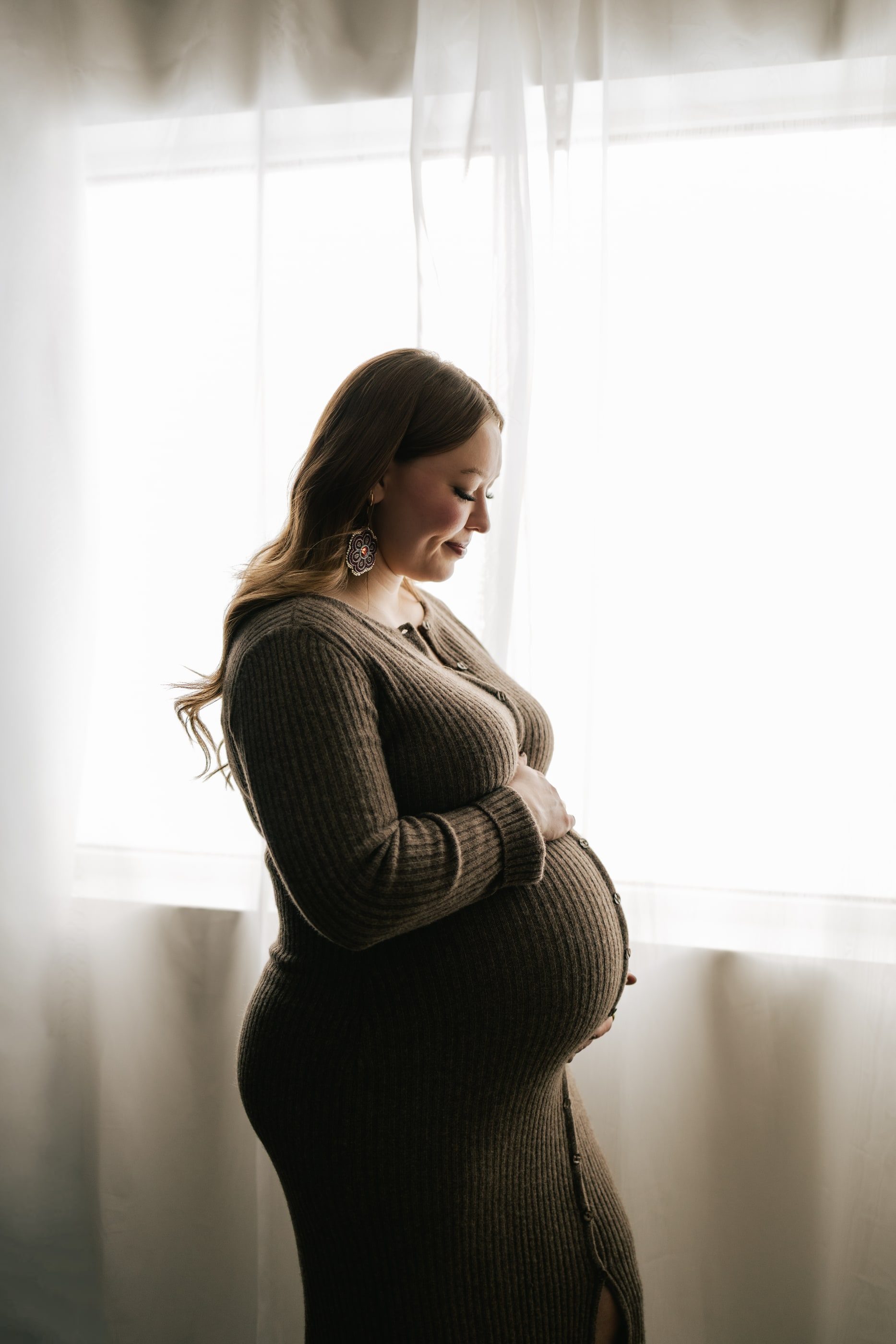 An expecting mother in a knit dress stands in profile by a sheer-curtained window, hands on her bump.