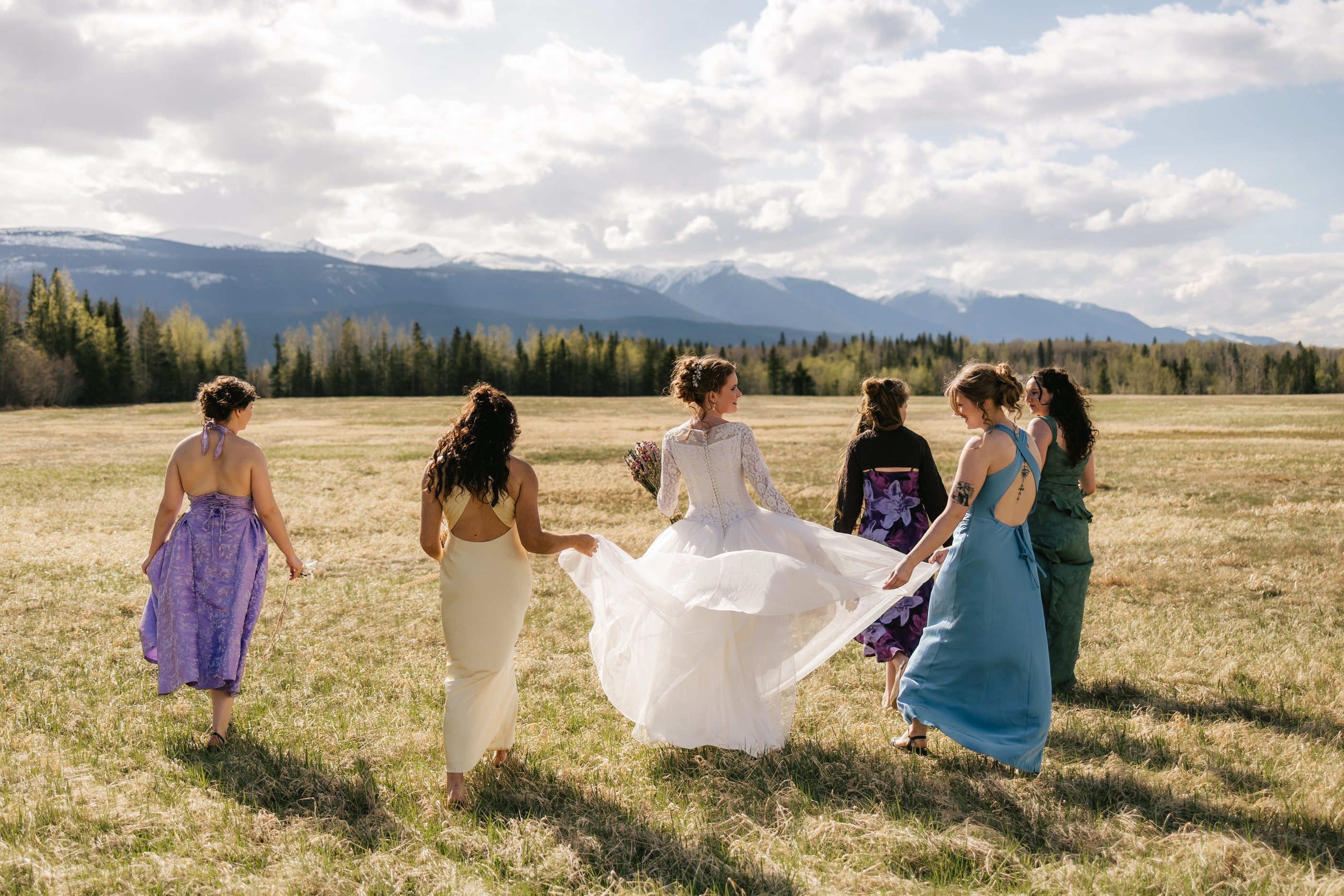 A bride and her bridesmaids in purple, gold, blue and green dresses walk through an open field toward distant mountains.
