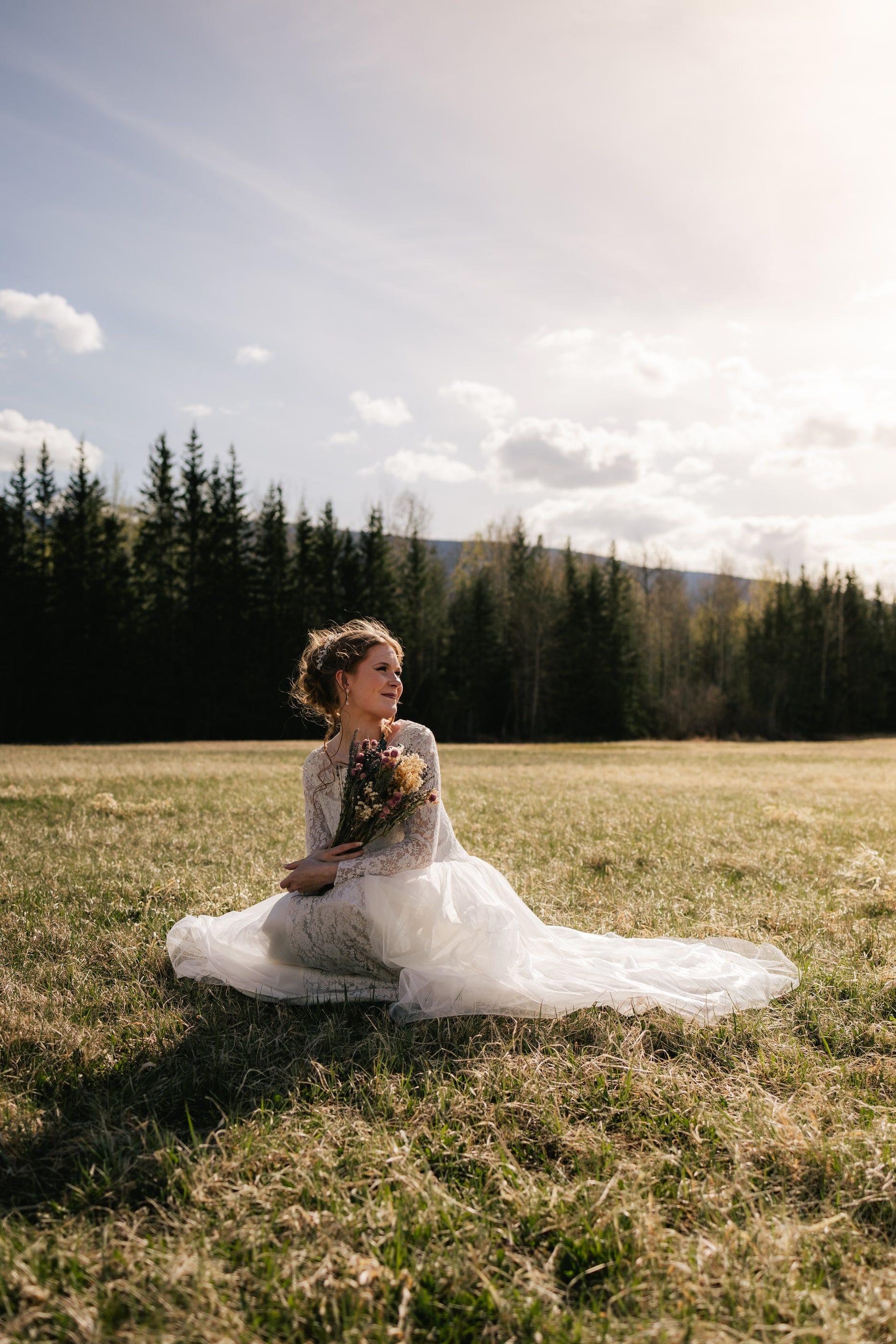 A bride sits in the grass of a sunlit meadow holding her bouquet, evergreens behind her.
