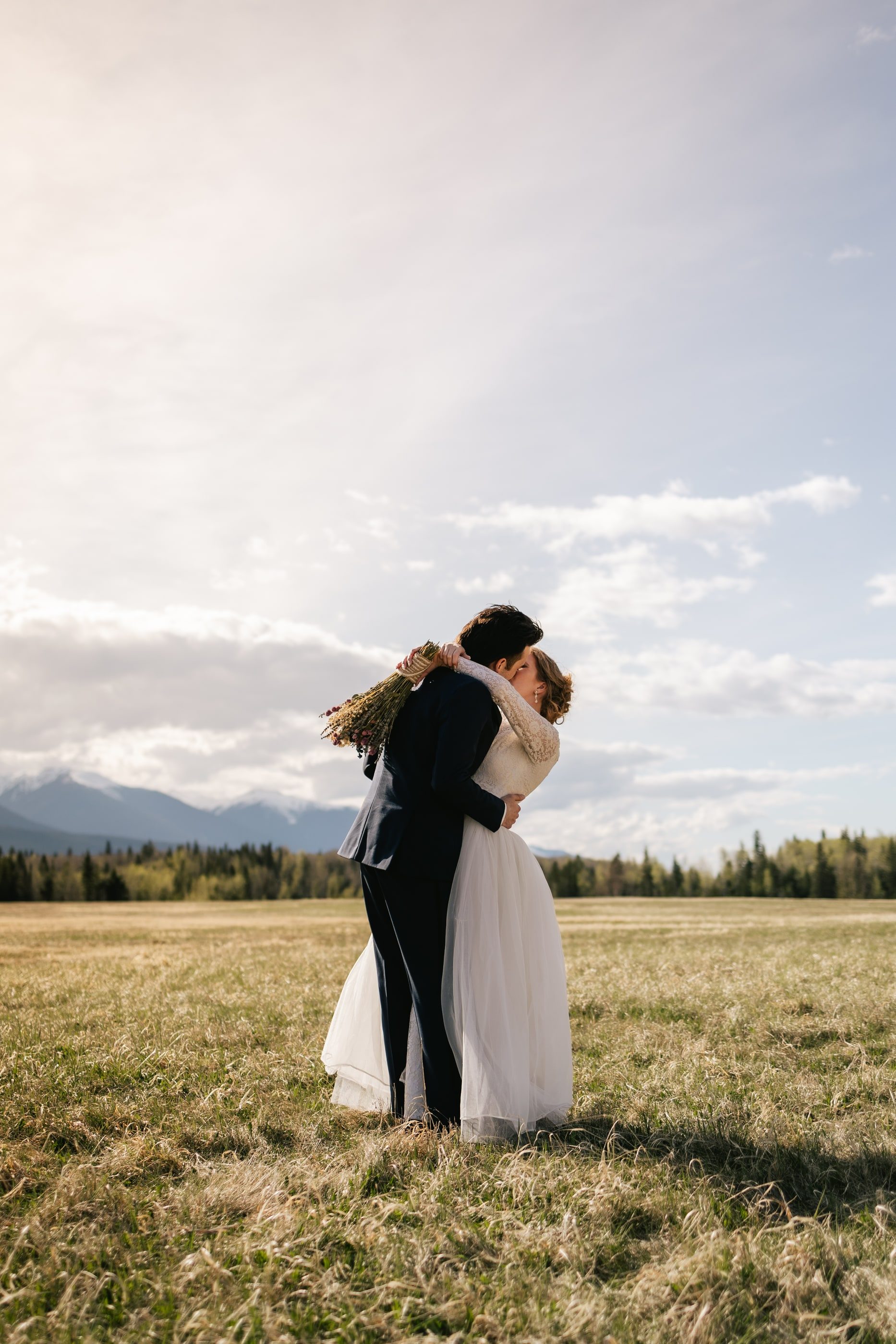 A bride and groom embrace in an open field beneath a bright sky, mountains on the horizon.