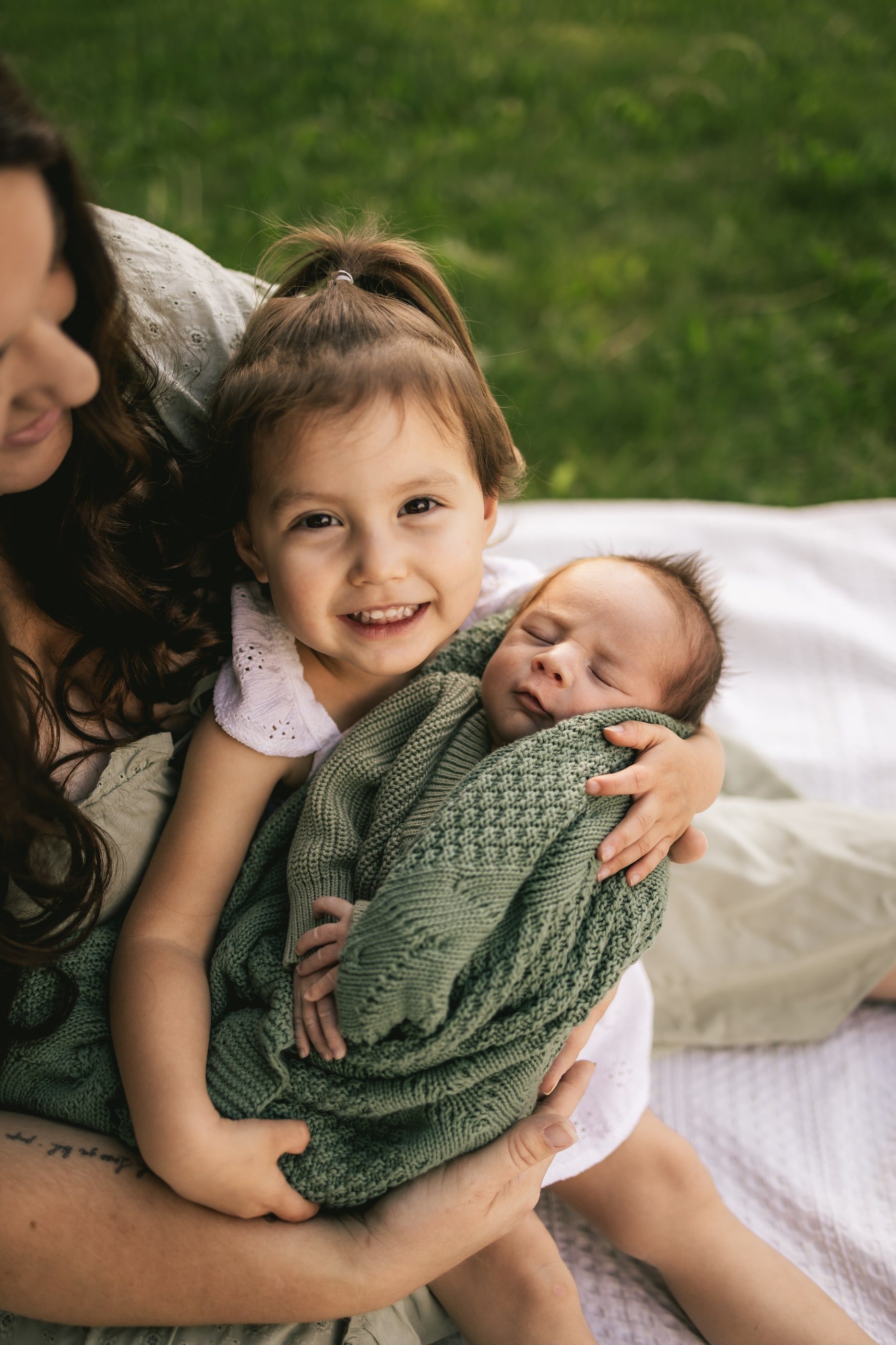 A smiling little girl holds her newborn sibling, wrapped in a green knit blanket, with her mother close beside them.