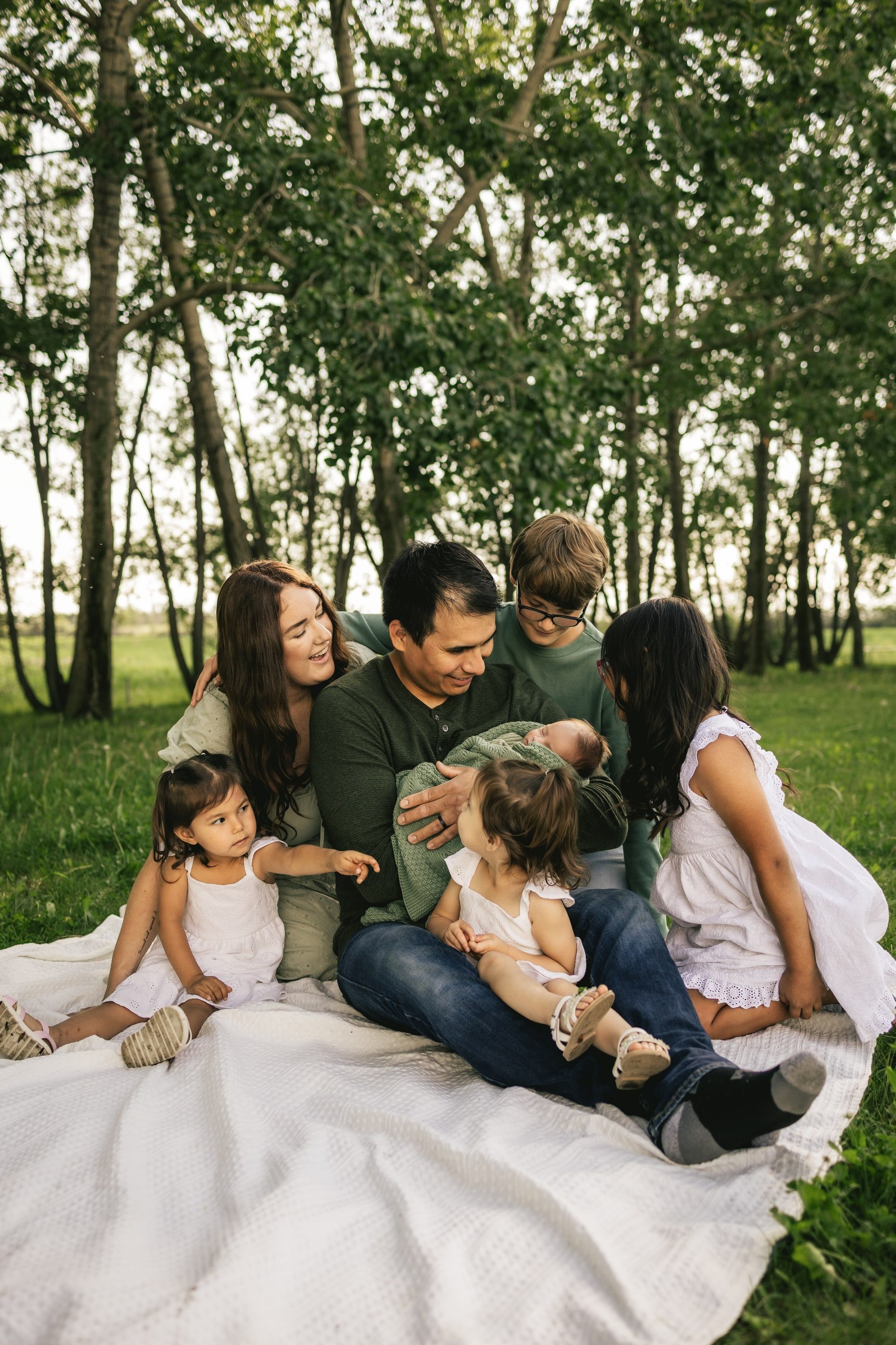 A family of five sits together on a white blanket in a grove of trees, the children laughing.