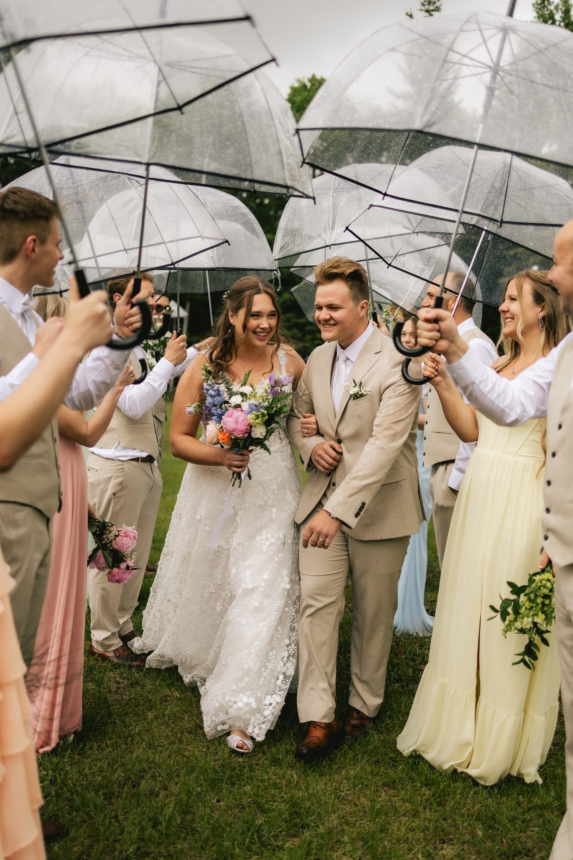 A bride and groom laugh as guests hold clear umbrellas over them on the lawn.