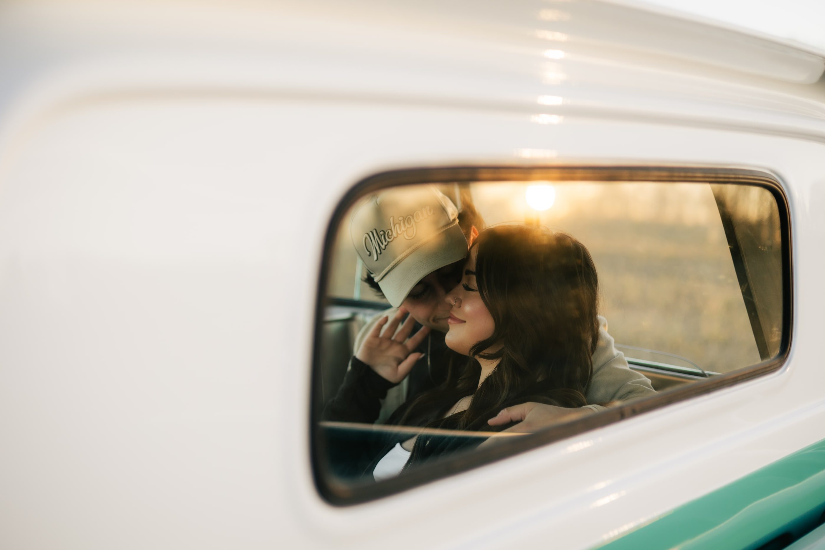 A couple embracing, framed through the side window of a vintage white truck at sunset.
