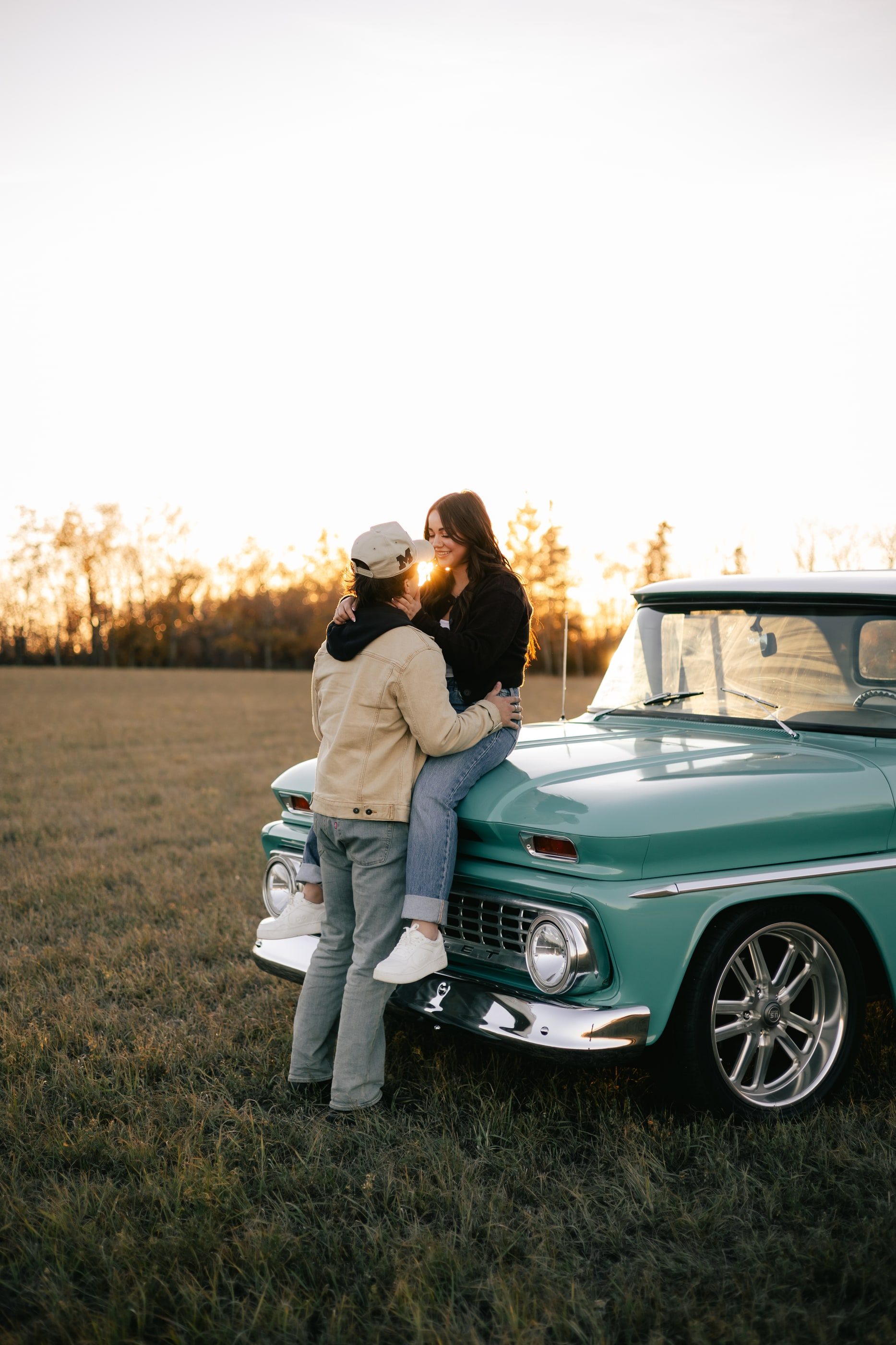 A woman sits on the hood of a vintage teal pickup with her arms around her partner, in a field at sunset.