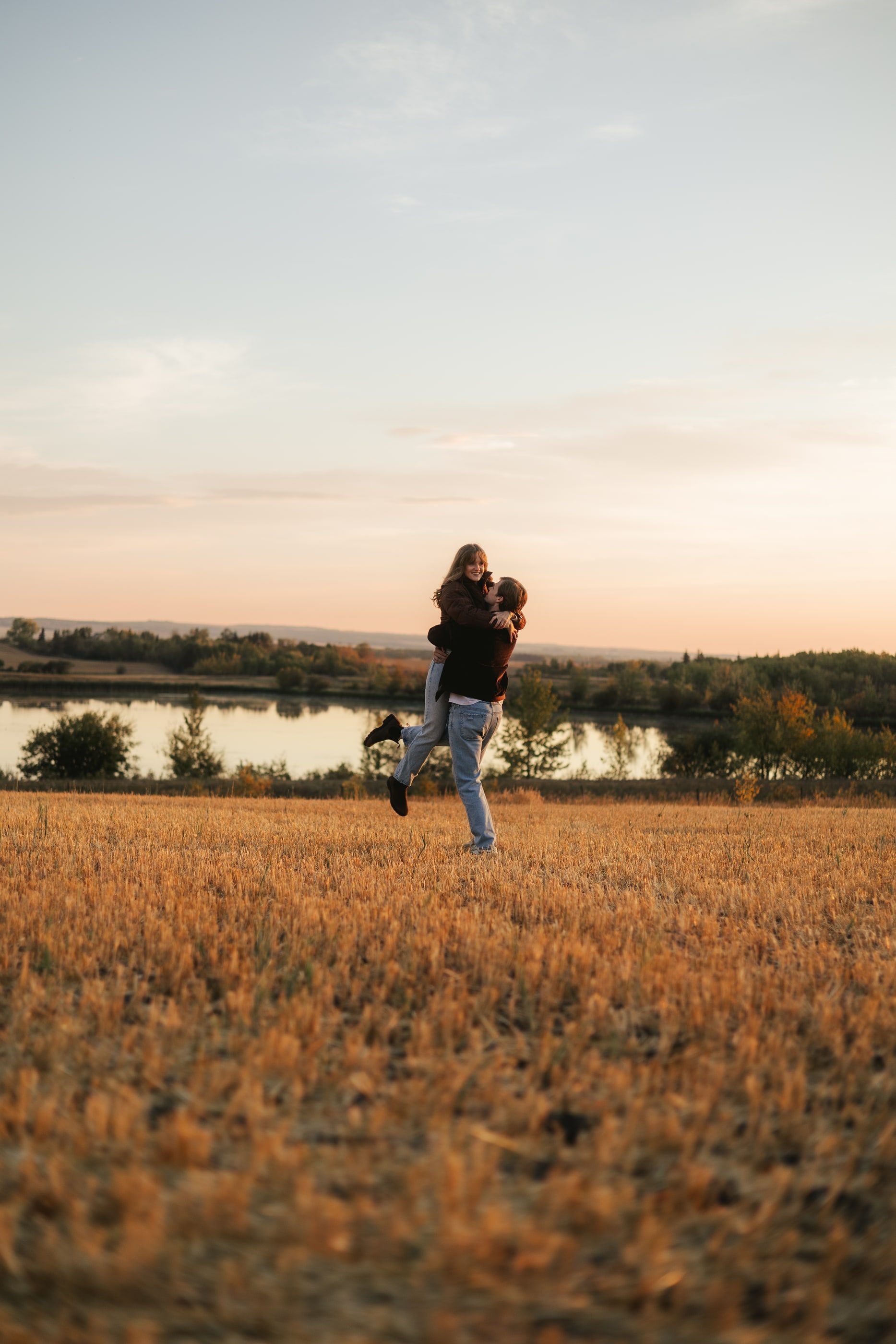 A man lifts his partner in a hug in a golden field beside a lake at dusk.