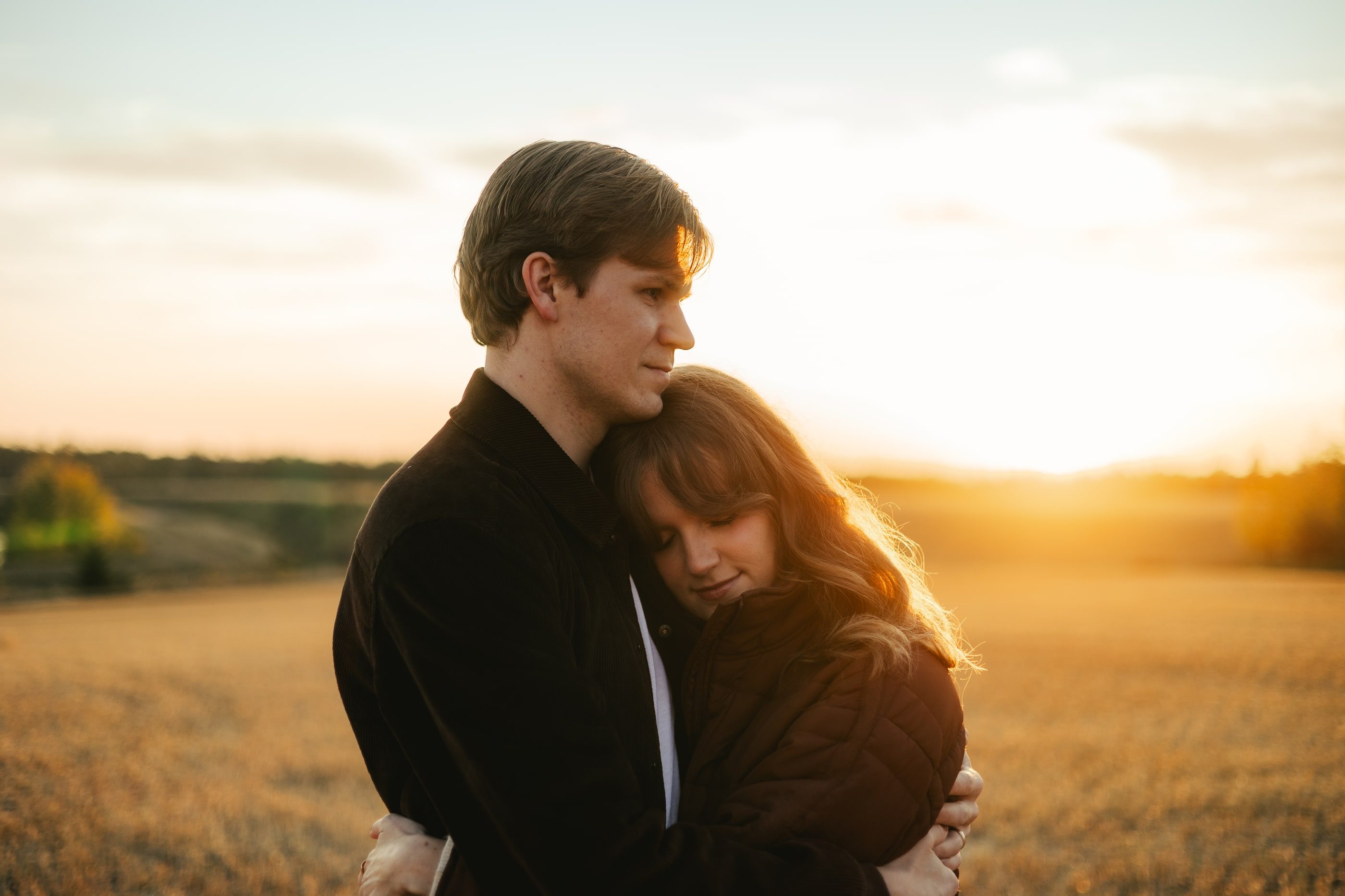A woman rests her head on her partner's chest as they stand in a field at golden hour.