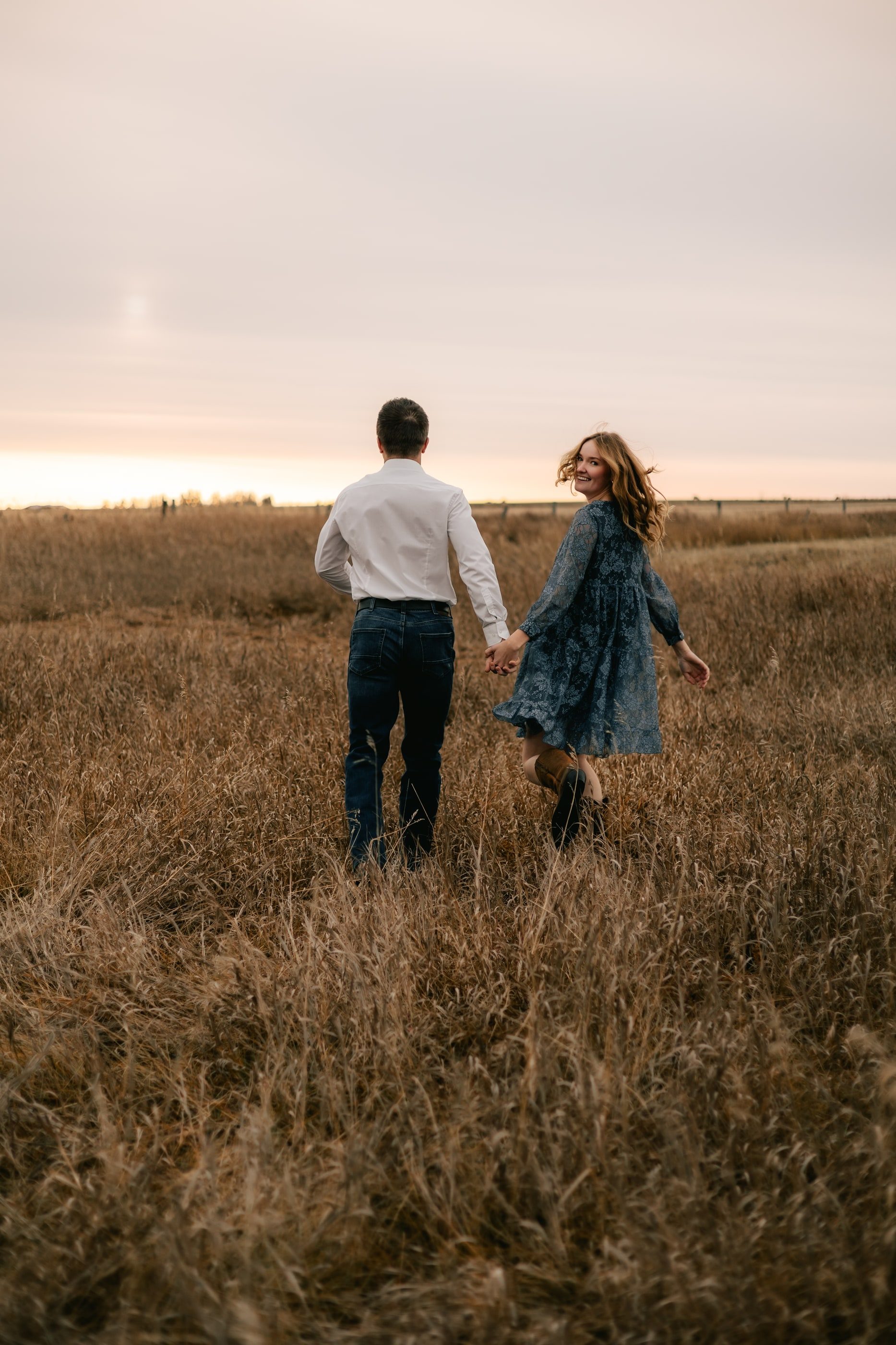 A couple walks hand in hand through tall prairie grass at dusk, she in a blue dress.