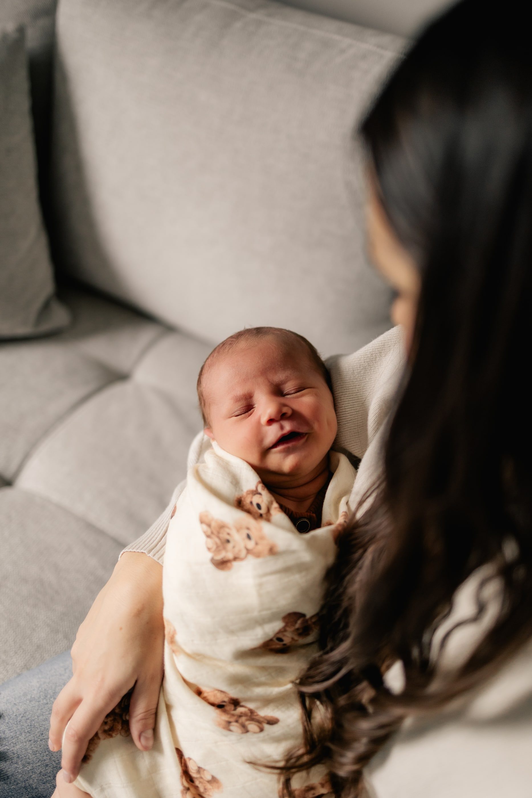 A mother cradles her newborn, swaddled in a teddy-bear print blanket, on a light grey sofa.