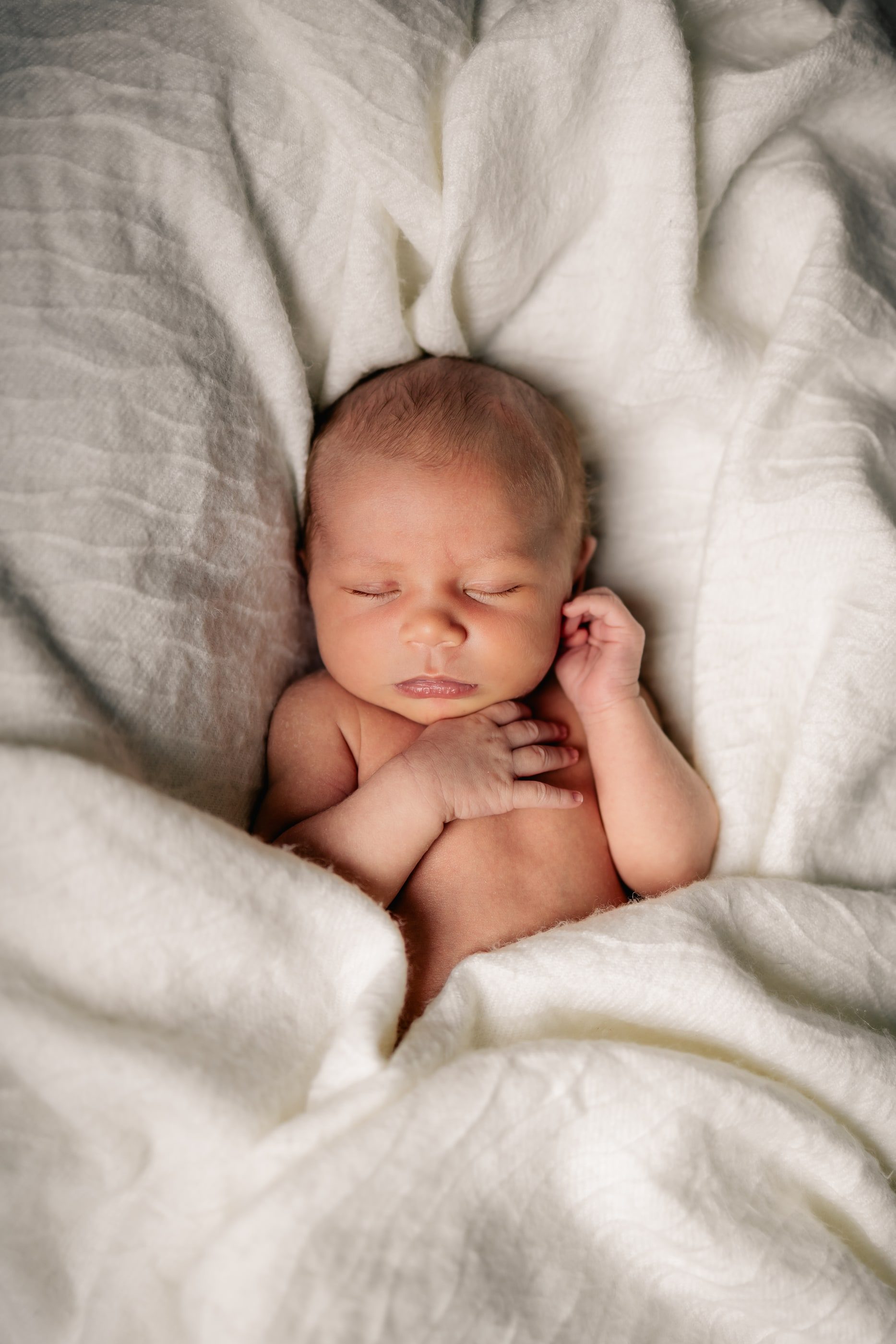 A sleeping newborn tucked into soft white linen, one hand by their cheek.