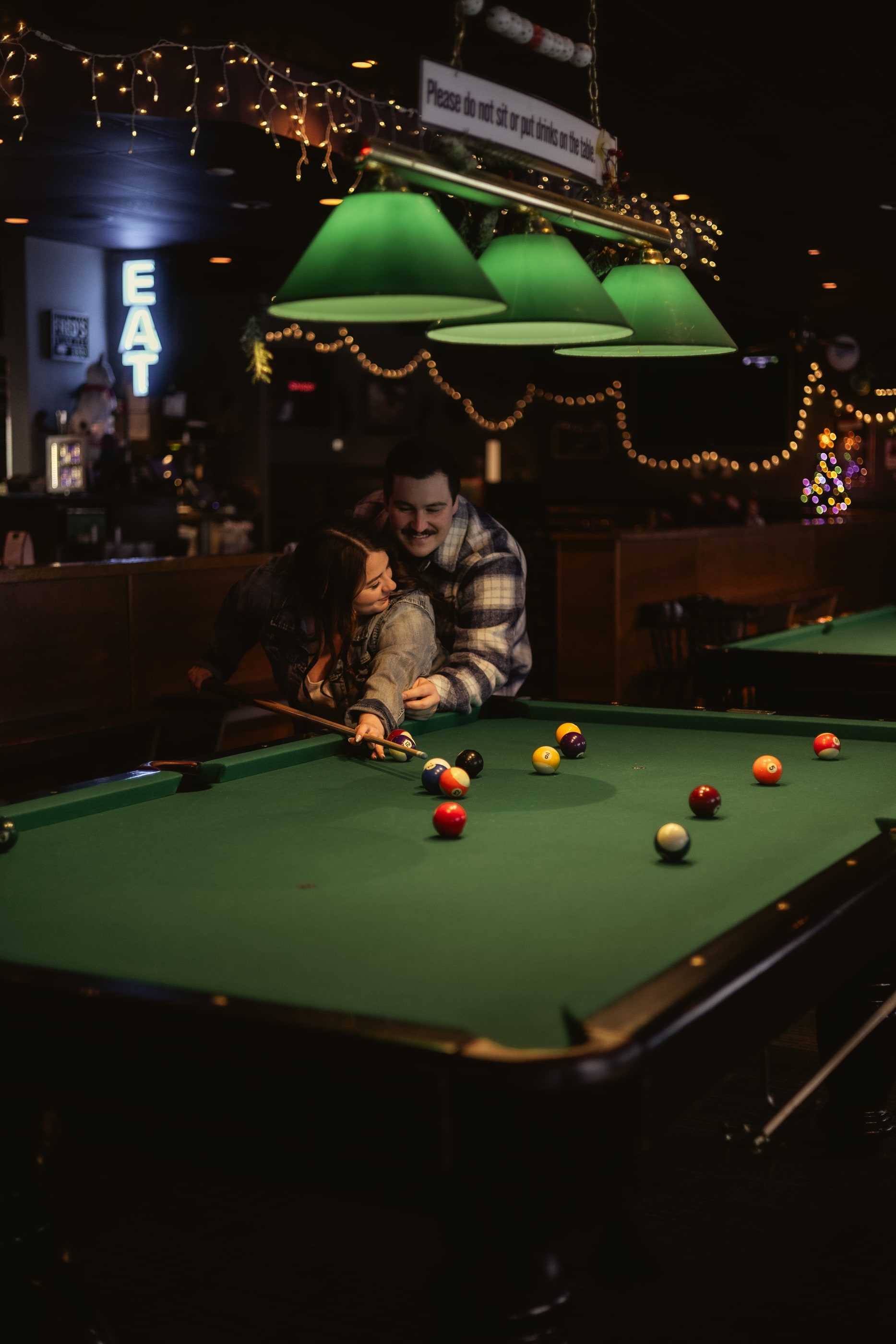 A man leans over a pool table to help his partner line up a shot in a dimly lit bar.