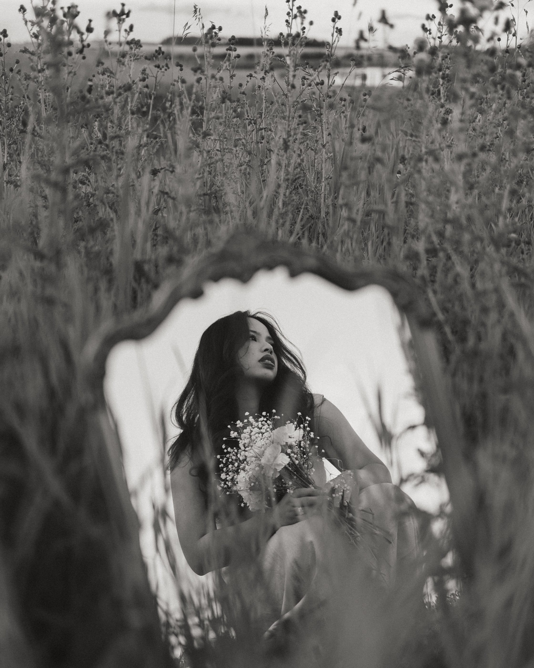 Black-and-white photo of a woman holding baby's breath, reflected in an ornate mirror set in tall grass.