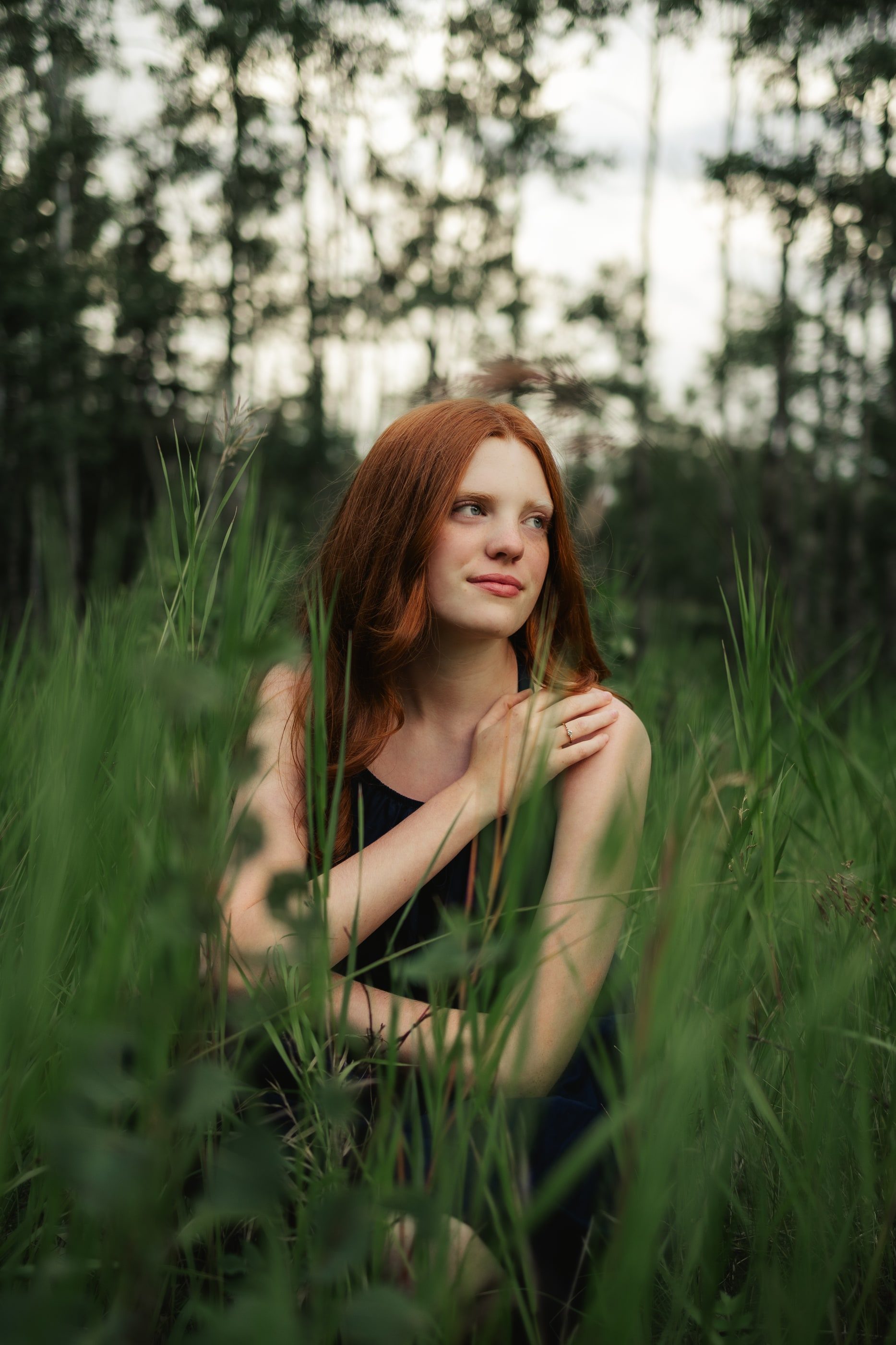A red-haired girl sits in tall green grass at the edge of the forest, smiling softly.