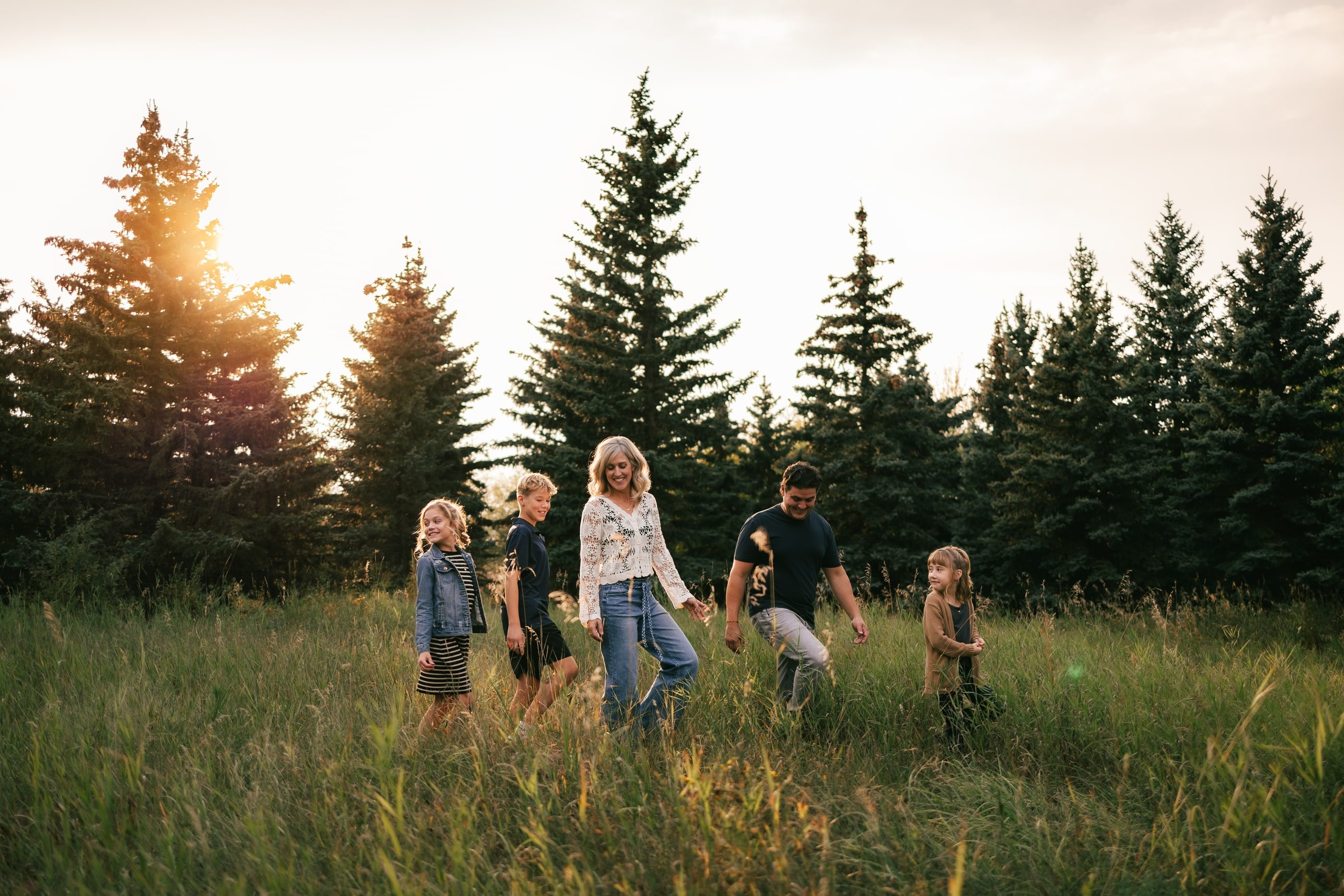 A family walks through a meadow lined with spruce trees as the sun sets.