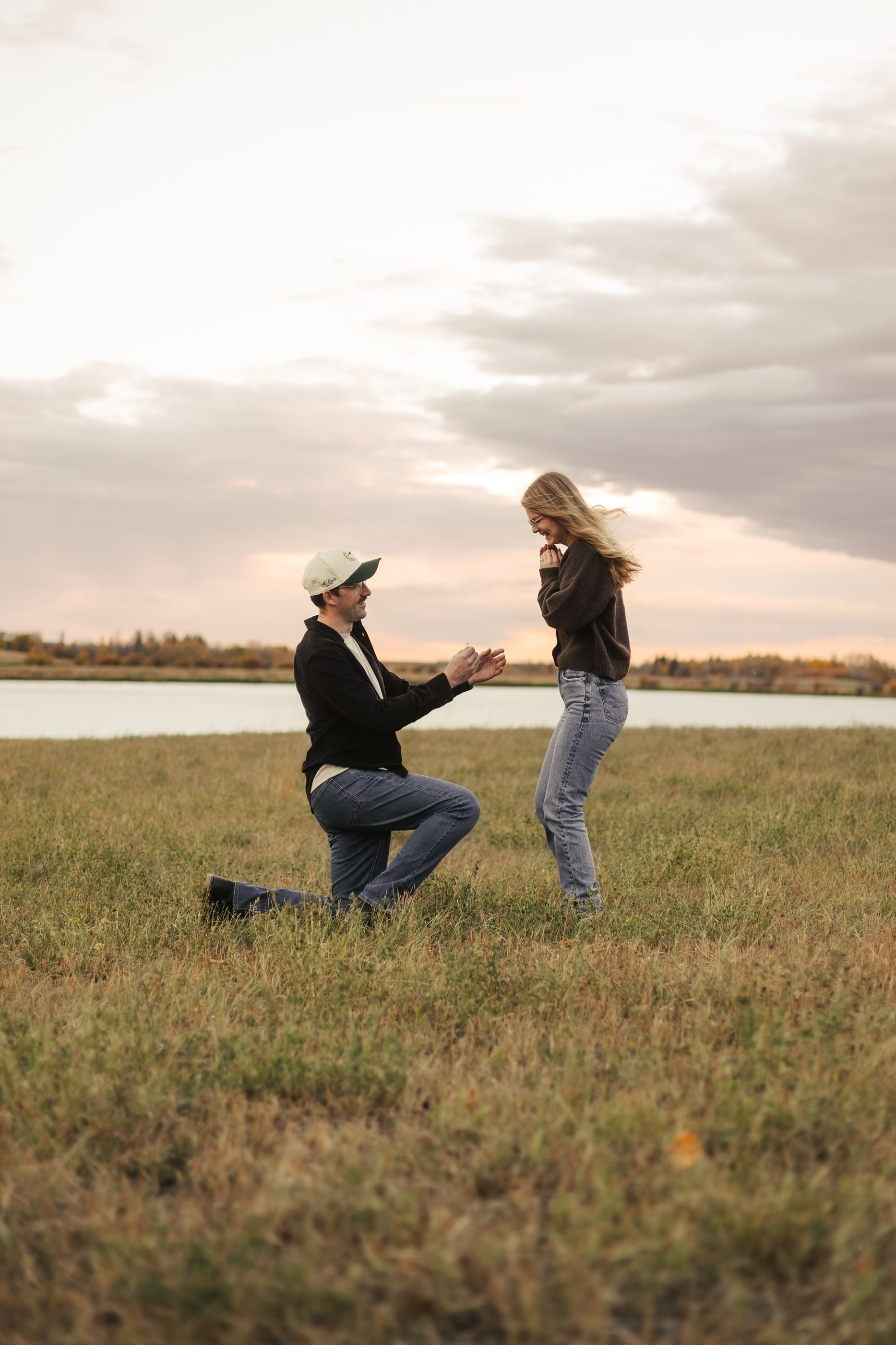 A man kneels to propose by a lake as his partner covers her mouth in surprise.