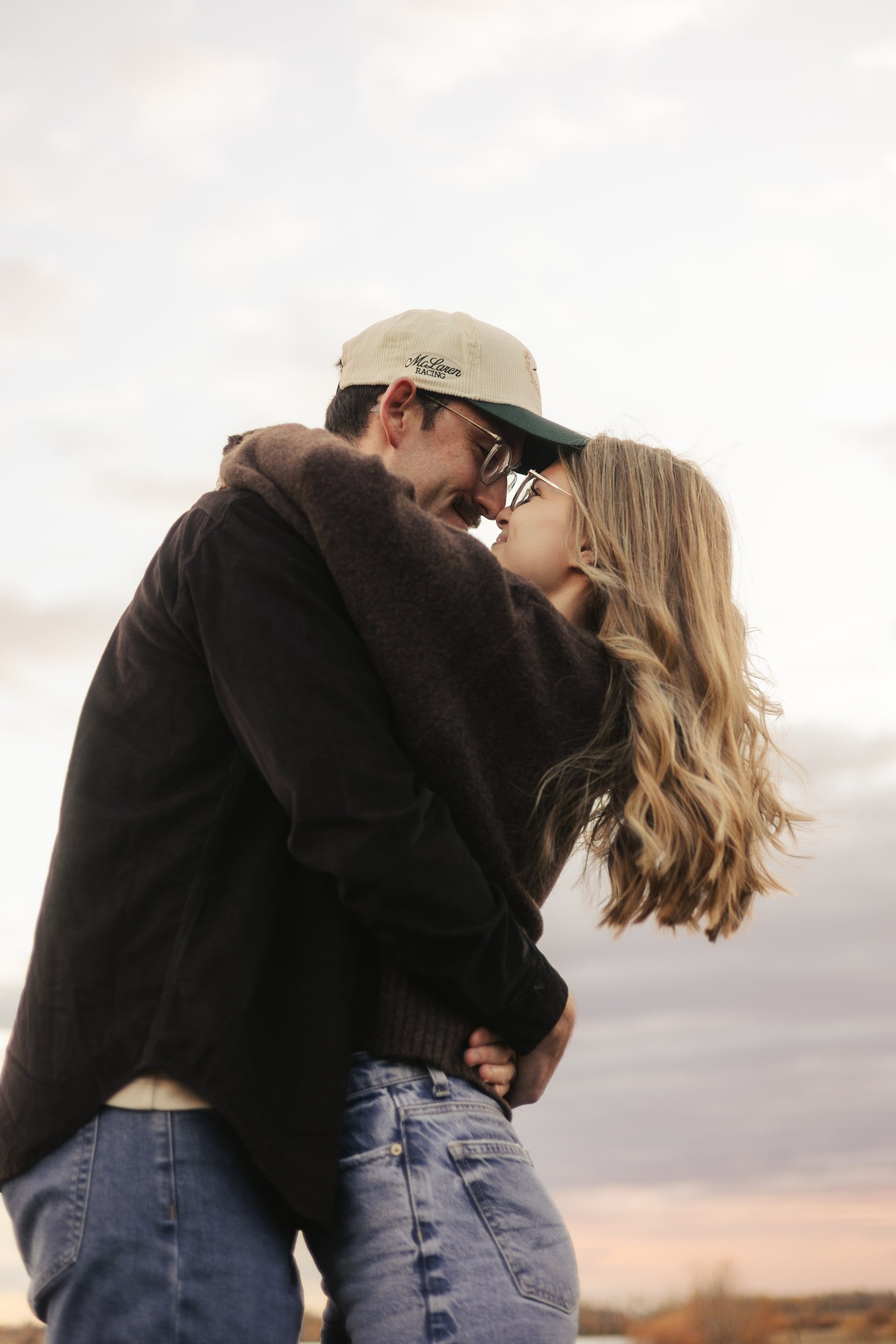 A couple holds each other close, her hair blowing in the wind, against a soft evening sky.