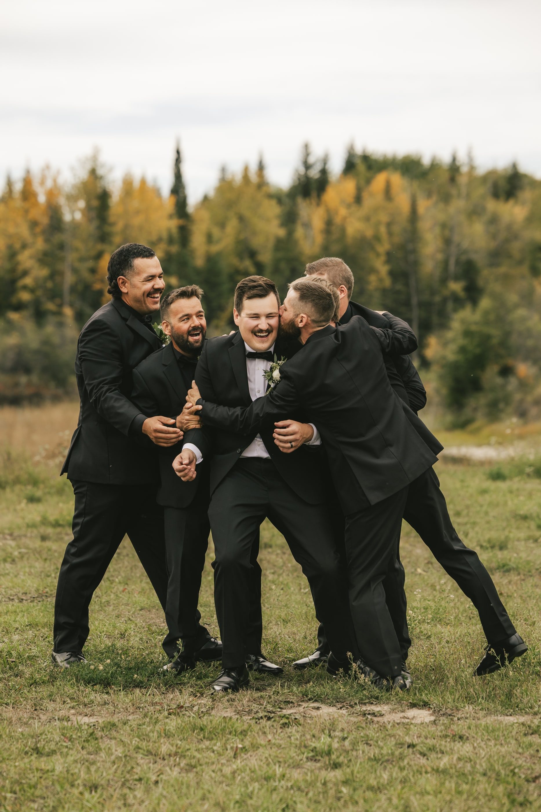 A groom laughs as his groomsmen in black suits pile in for a hug in an autumn field.