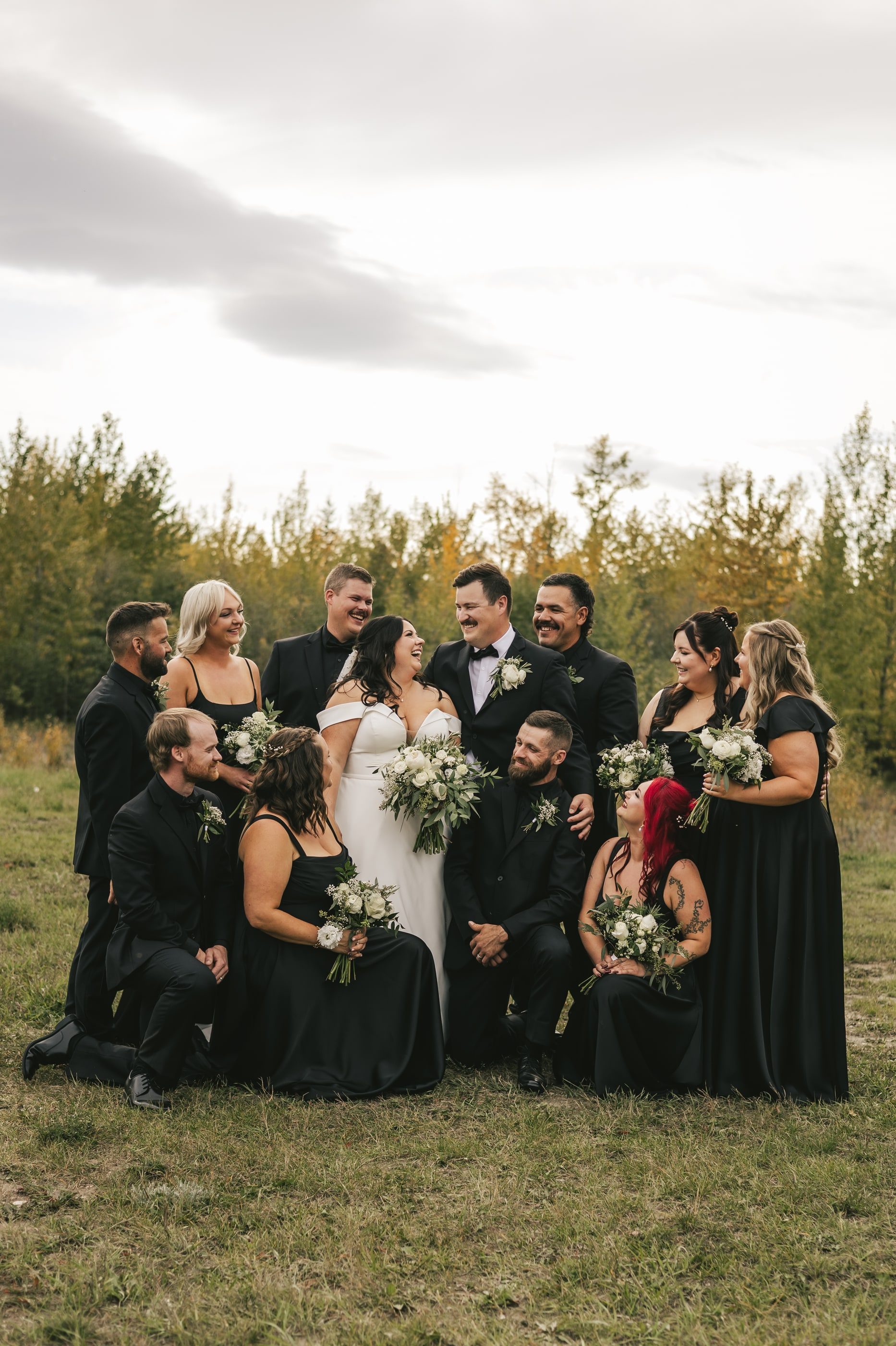 A wedding party in black gathers around the bride and groom in a field ringed by autumn trees.