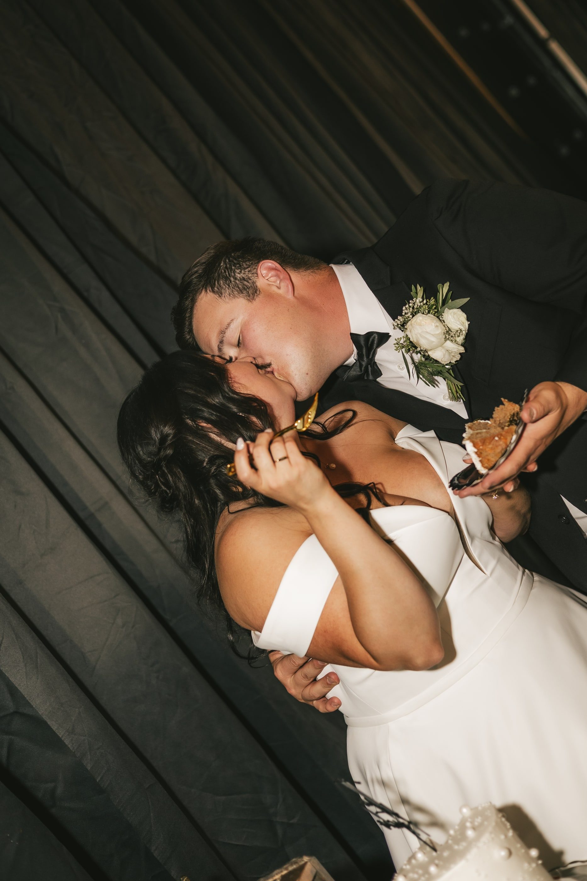 A groom kisses his laughing bride as they share a bite at the reception.
