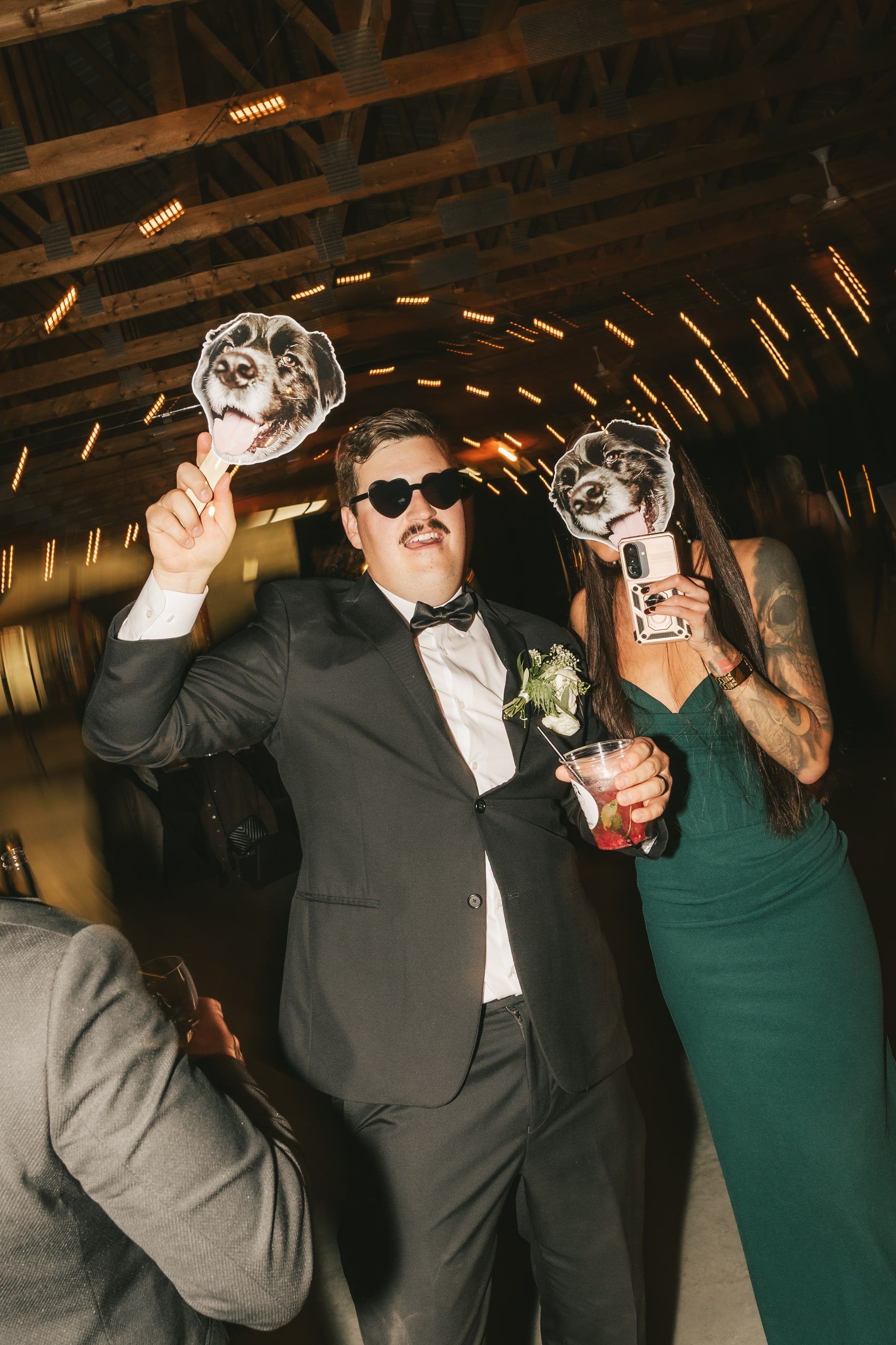 A groom in sunglasses dances with guests holding up photo cut-outs of faces on the reception dance floor.