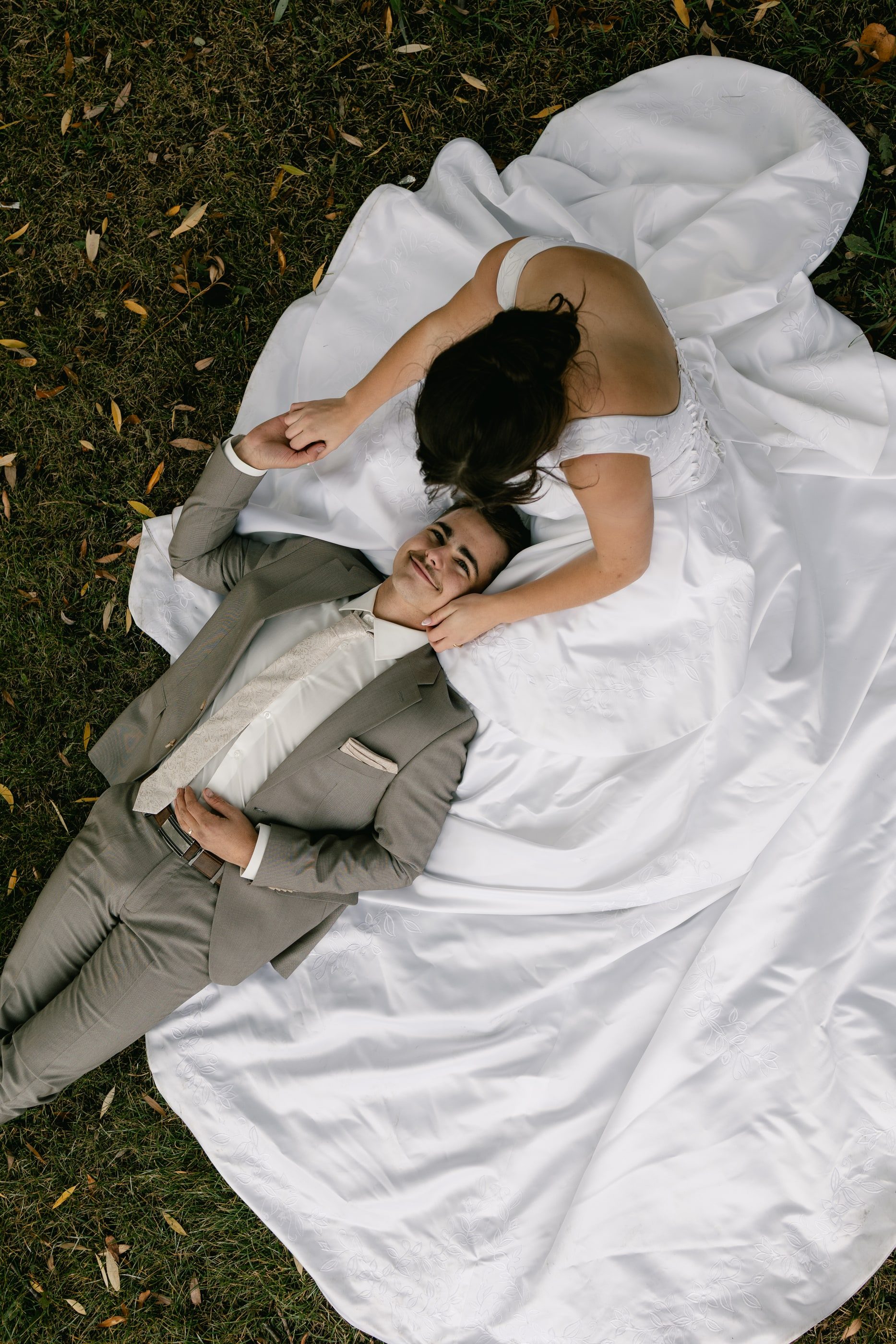 Seen from above, a bride and groom lie on the grass, her wide white gown spread around them.