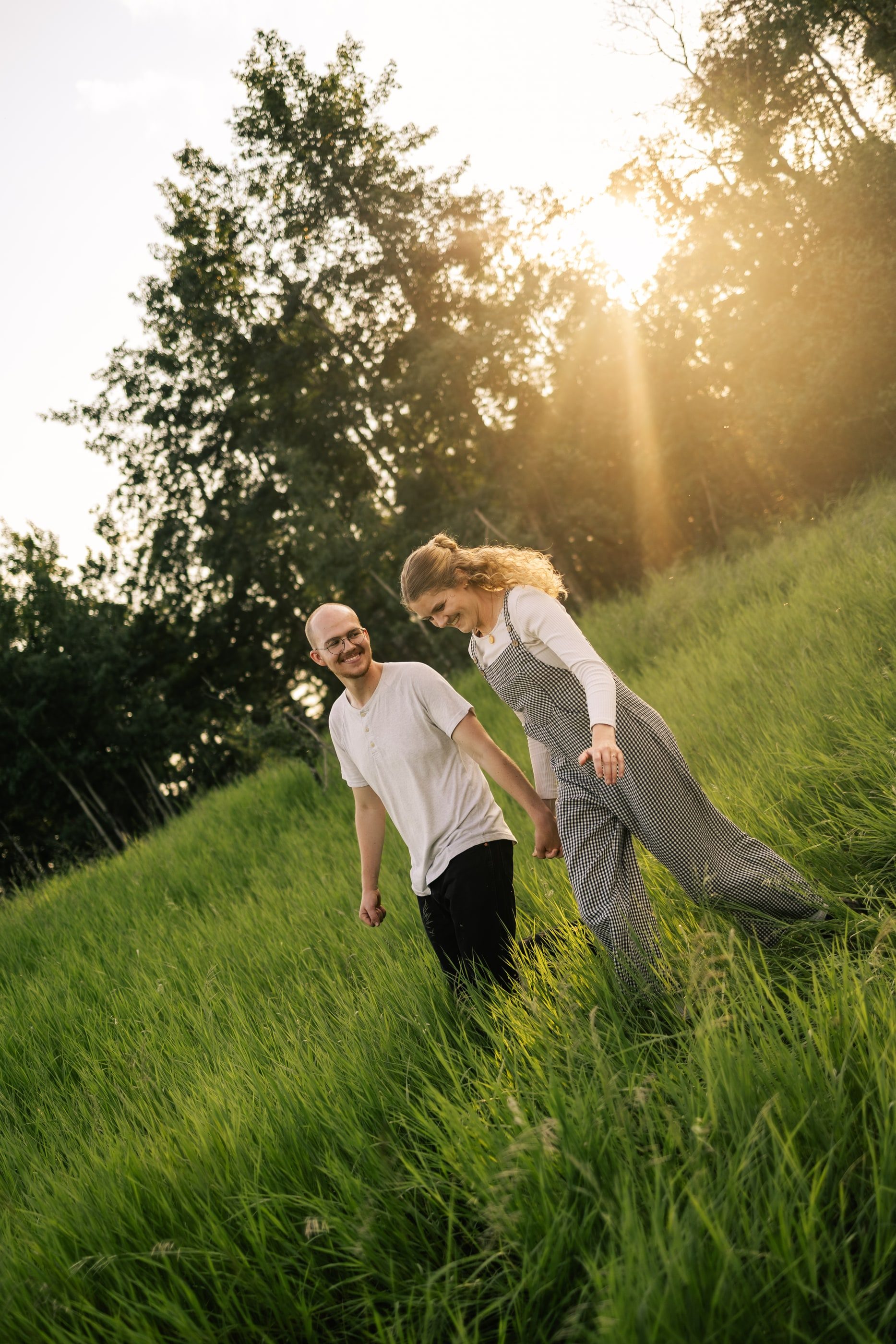A couple walks hand in hand down a grassy hill, sunlight streaming through the trees.