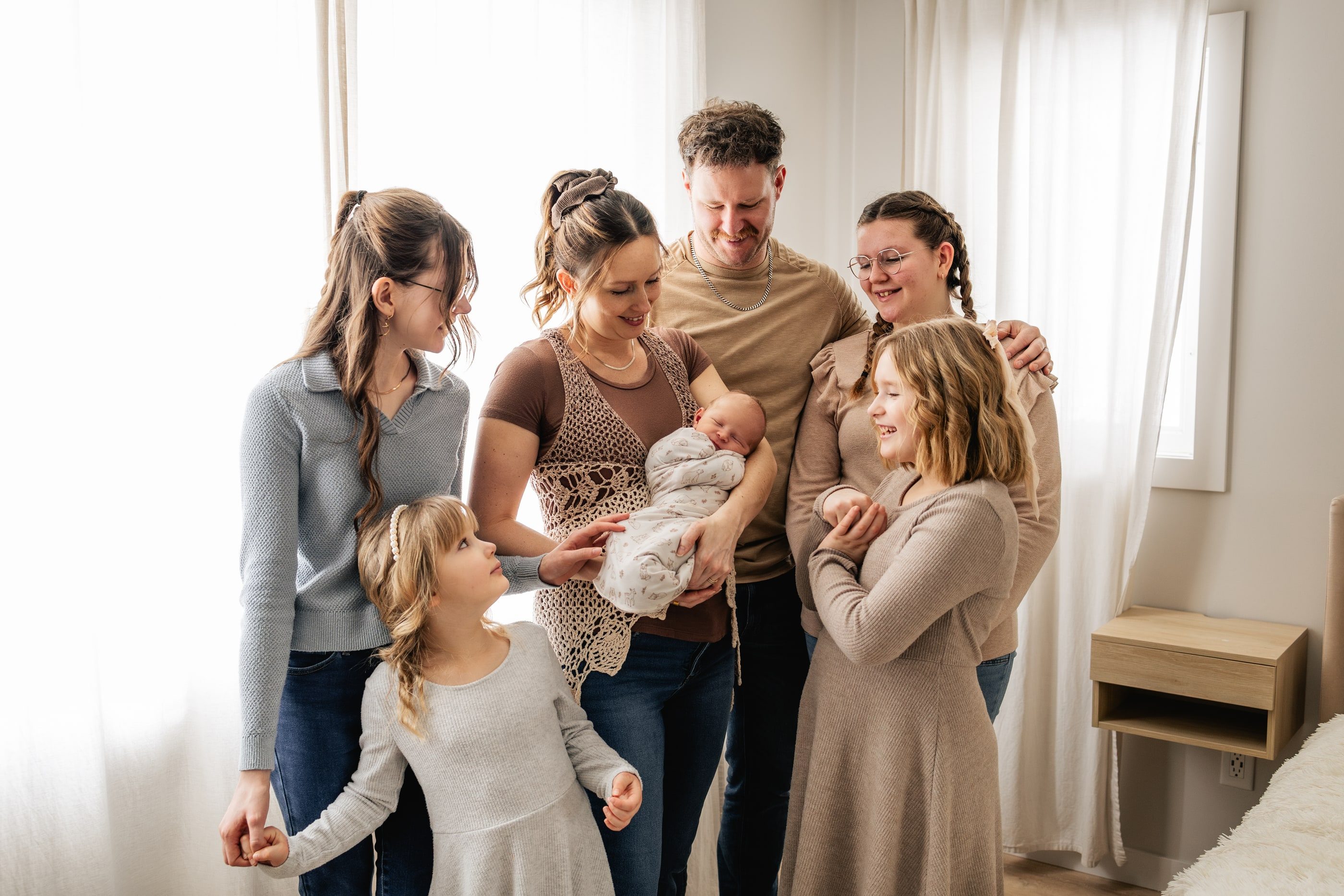 A family gathers around parents holding their newborn in a bright bedroom.