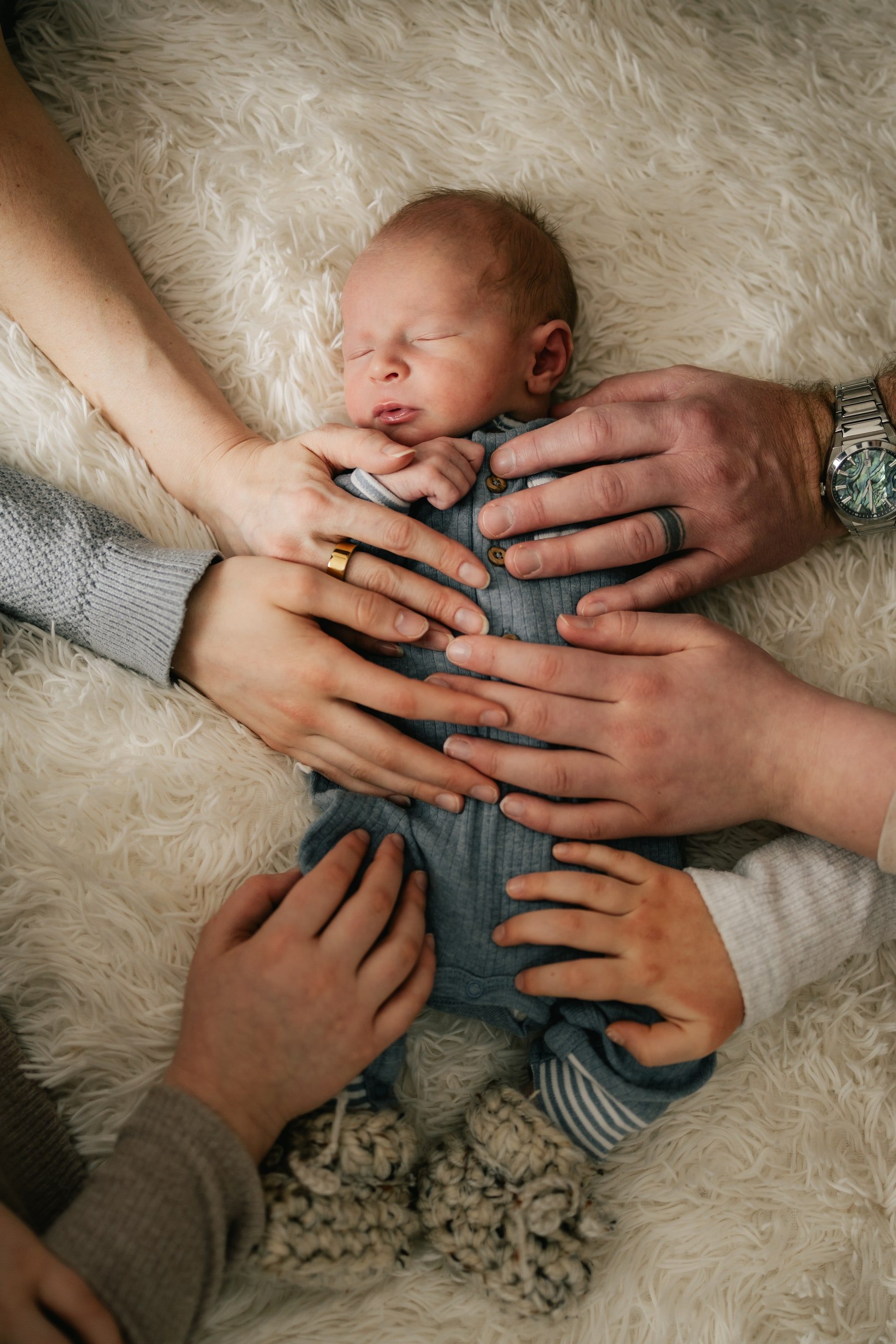 Hands from the whole family rest gently on a sleeping newborn on a sheepskin rug.