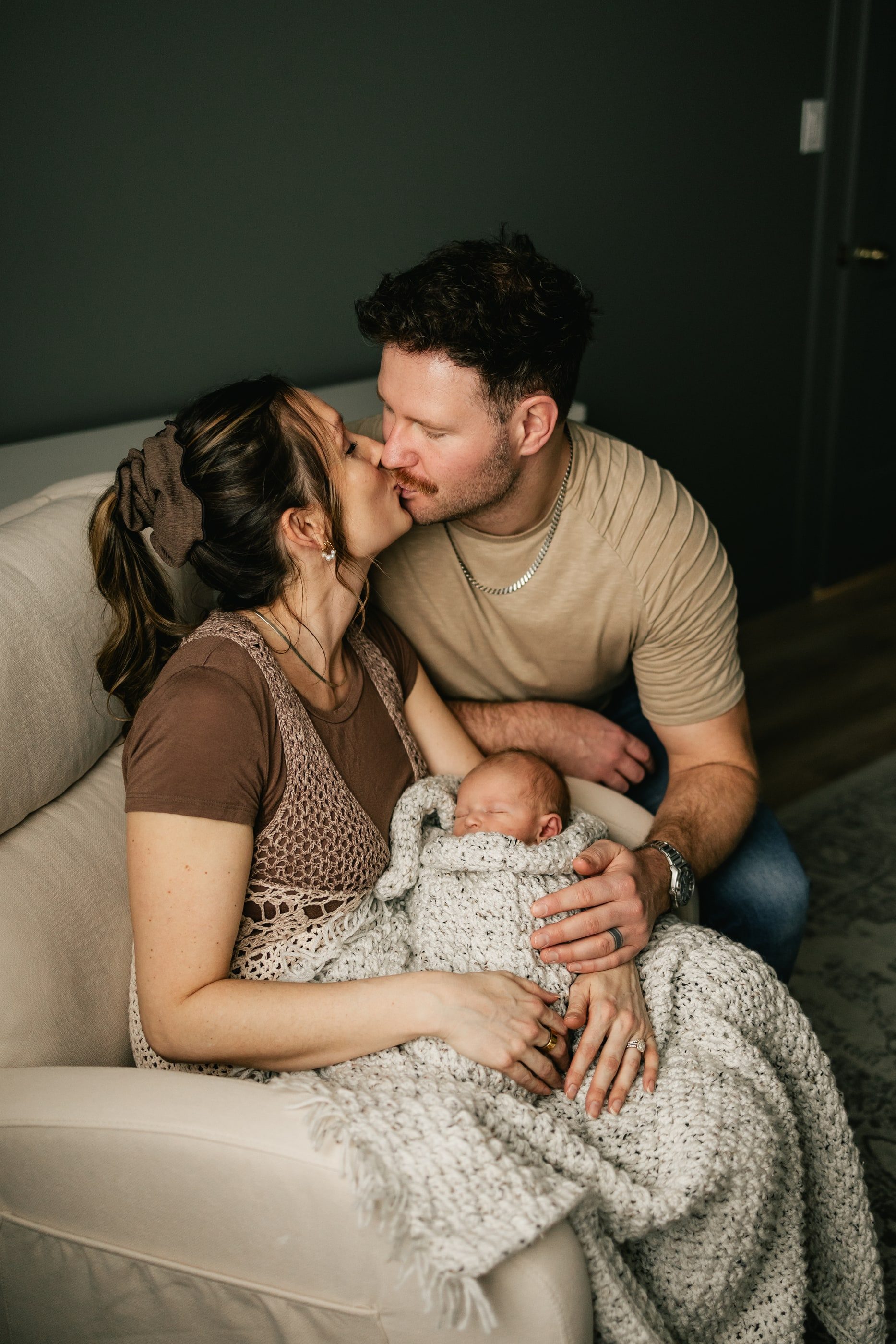 Parents kiss as they hold their newborn, wrapped in a knit blanket, in an armchair.