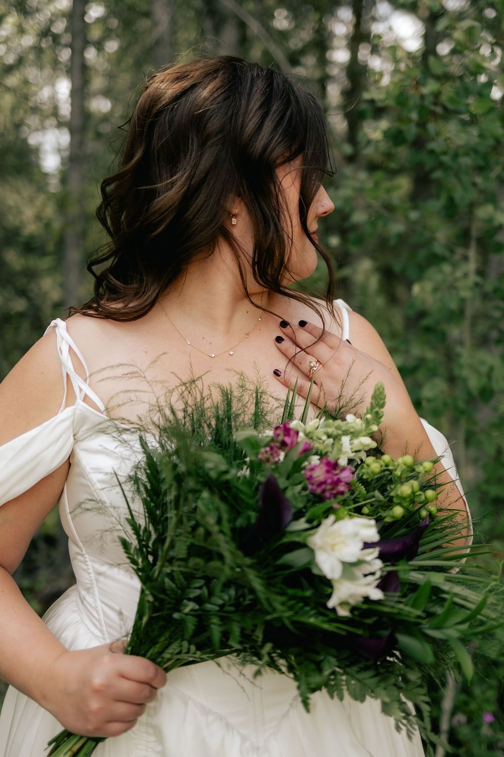 A bride glances over her shoulder, holding a bouquet of greenery and white flowers.