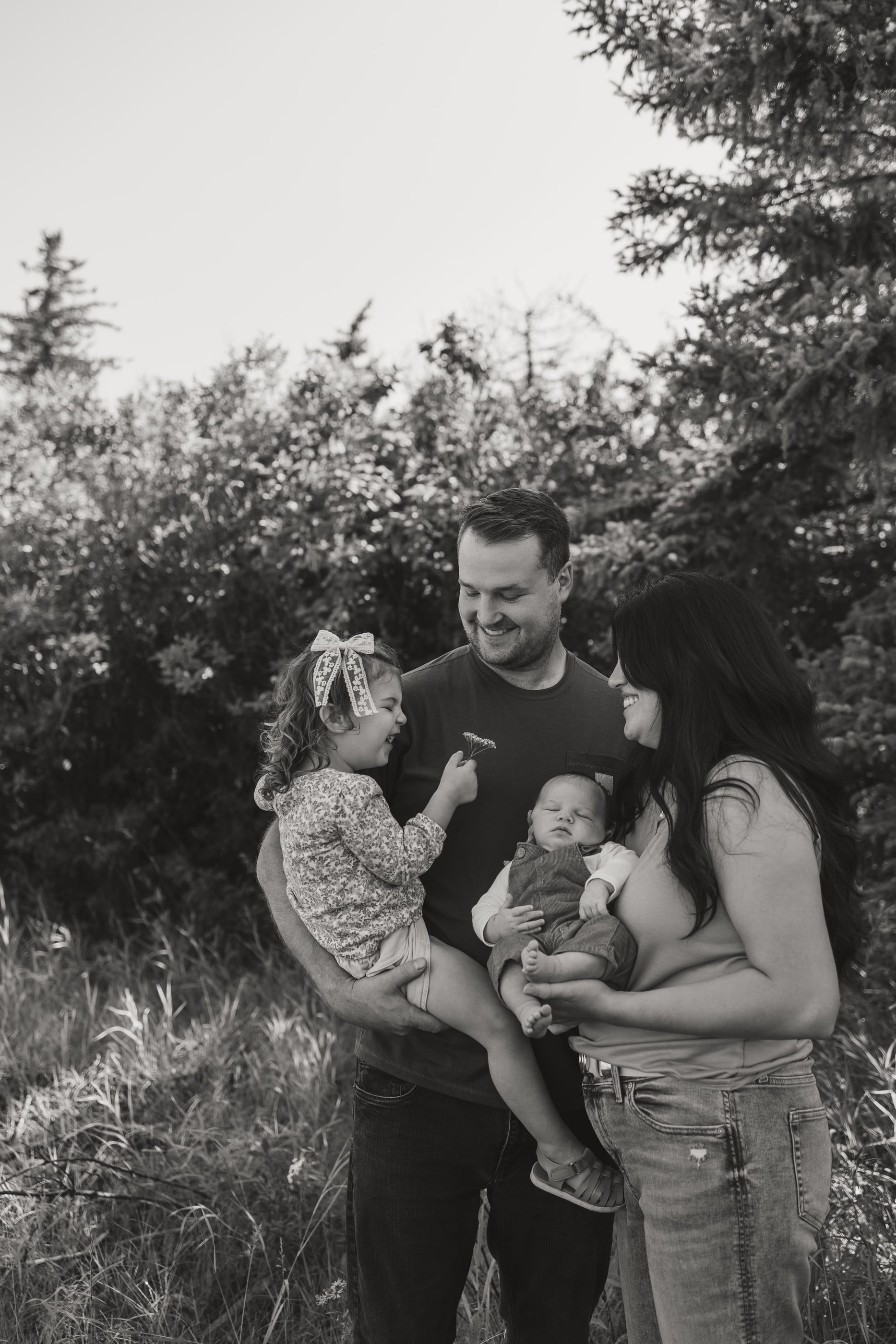 Black-and-white photo of parents with their toddler and newborn among evergreens.