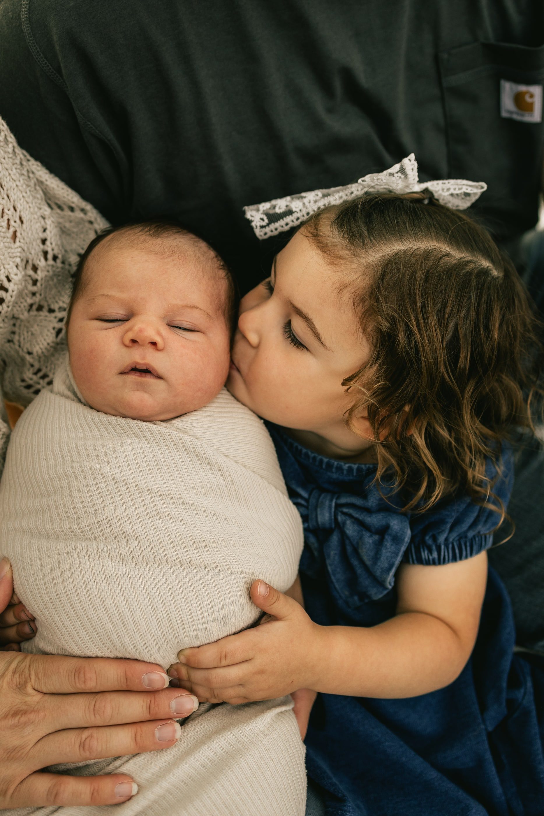 A little girl with a bow in her hair kisses her swaddled newborn sibling.