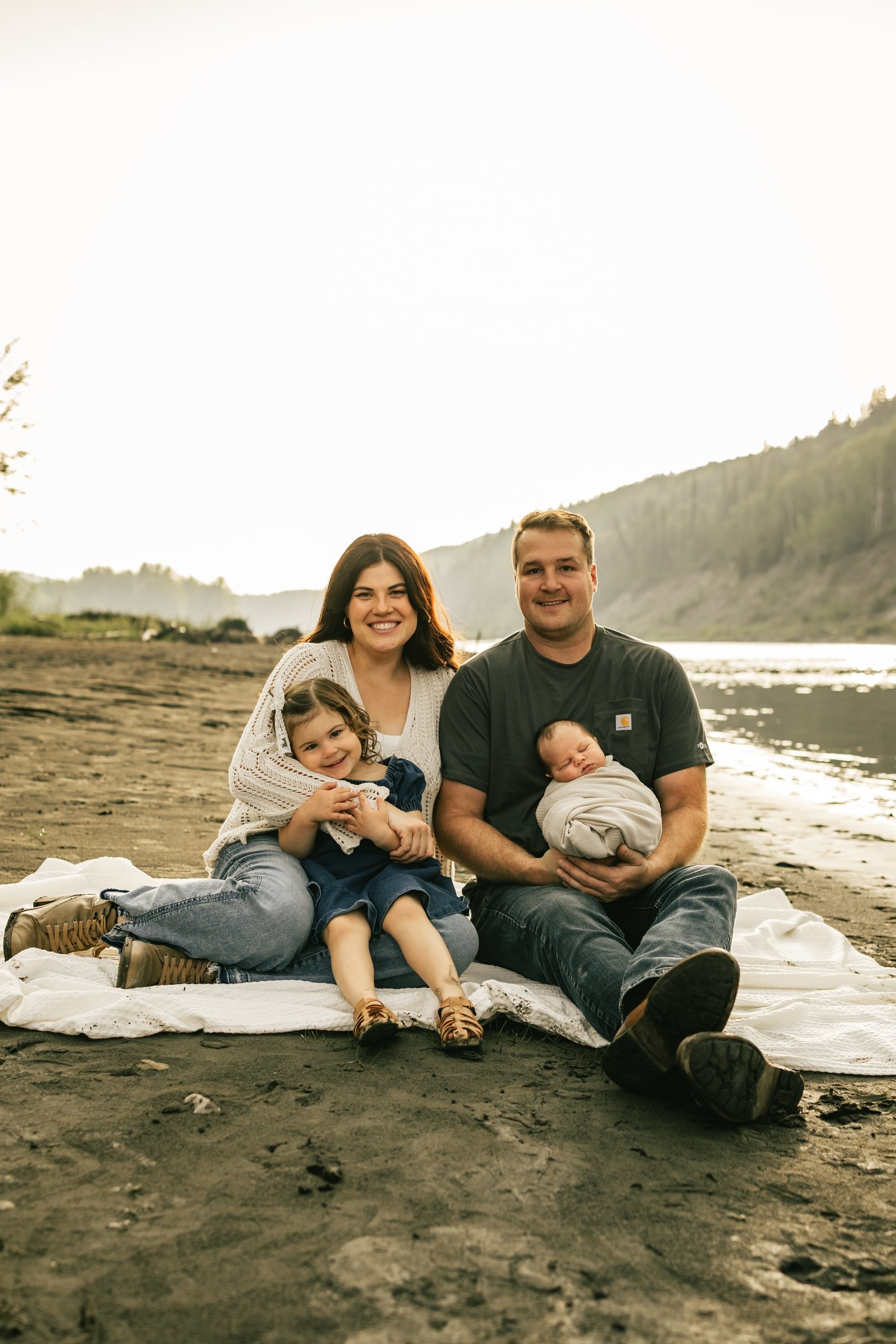 A family of four sits on a blanket on a riverbank, the father holding the newborn.