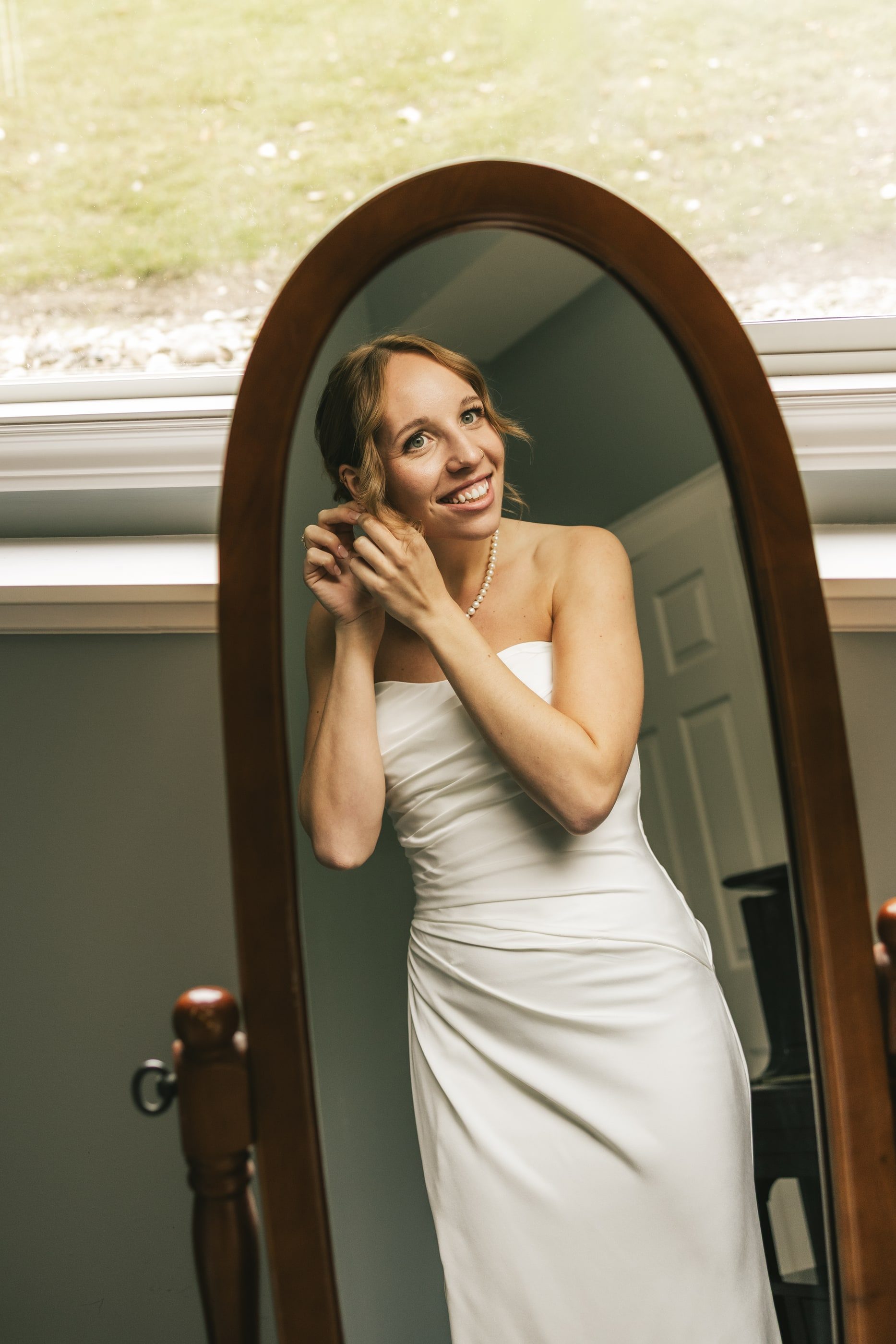 A bride smiles at her reflection in a tall wooden mirror as she fastens an earring.