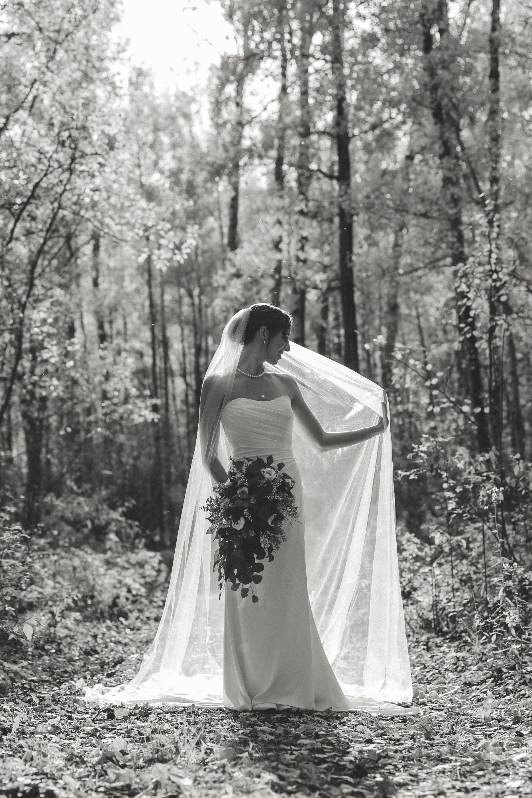Black-and-white photo of a bride in a long veil standing among birch trees, holding a cascading bouquet.