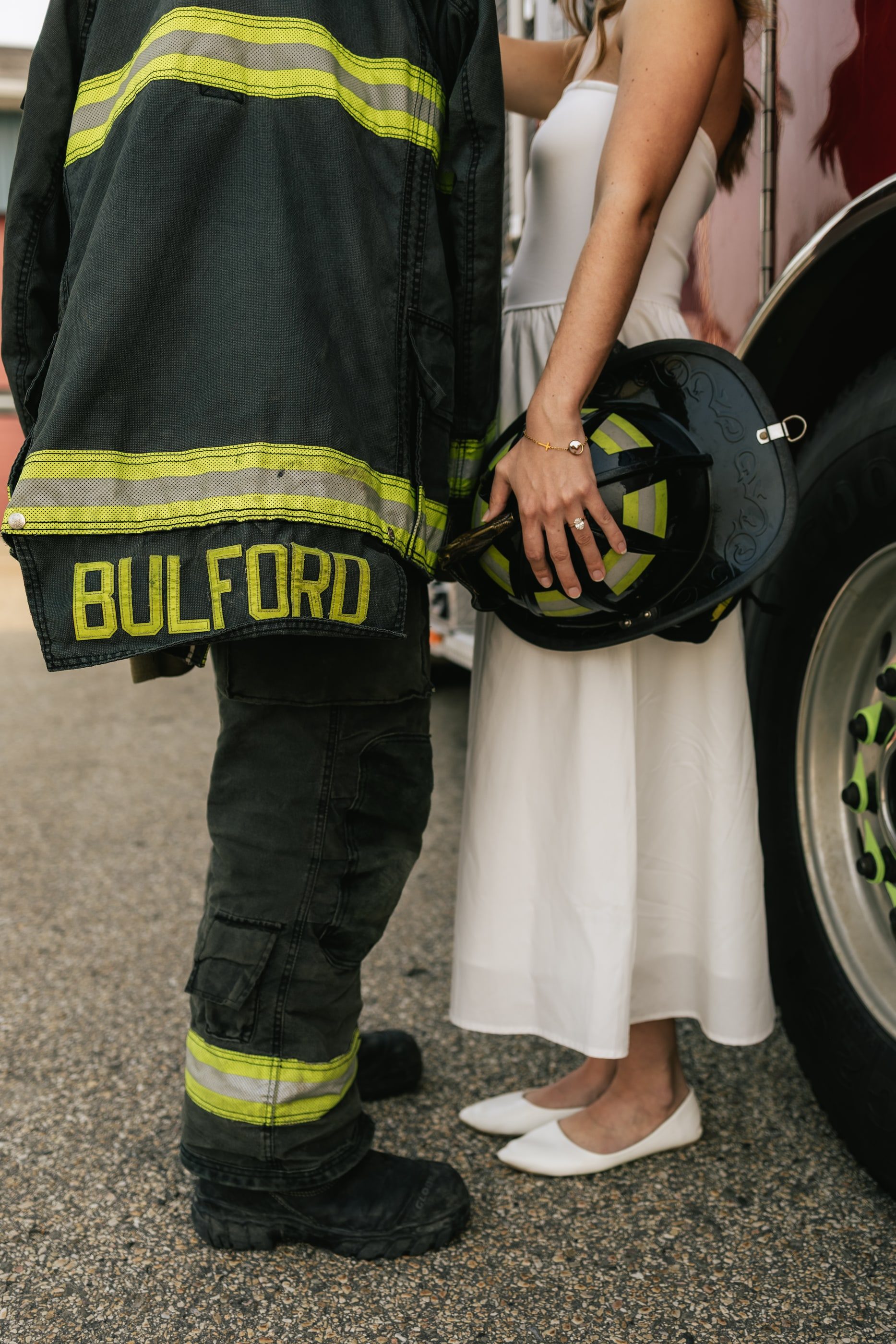A firefighter in turnout gear stands beside his partner in a white dress, who holds his helmet.