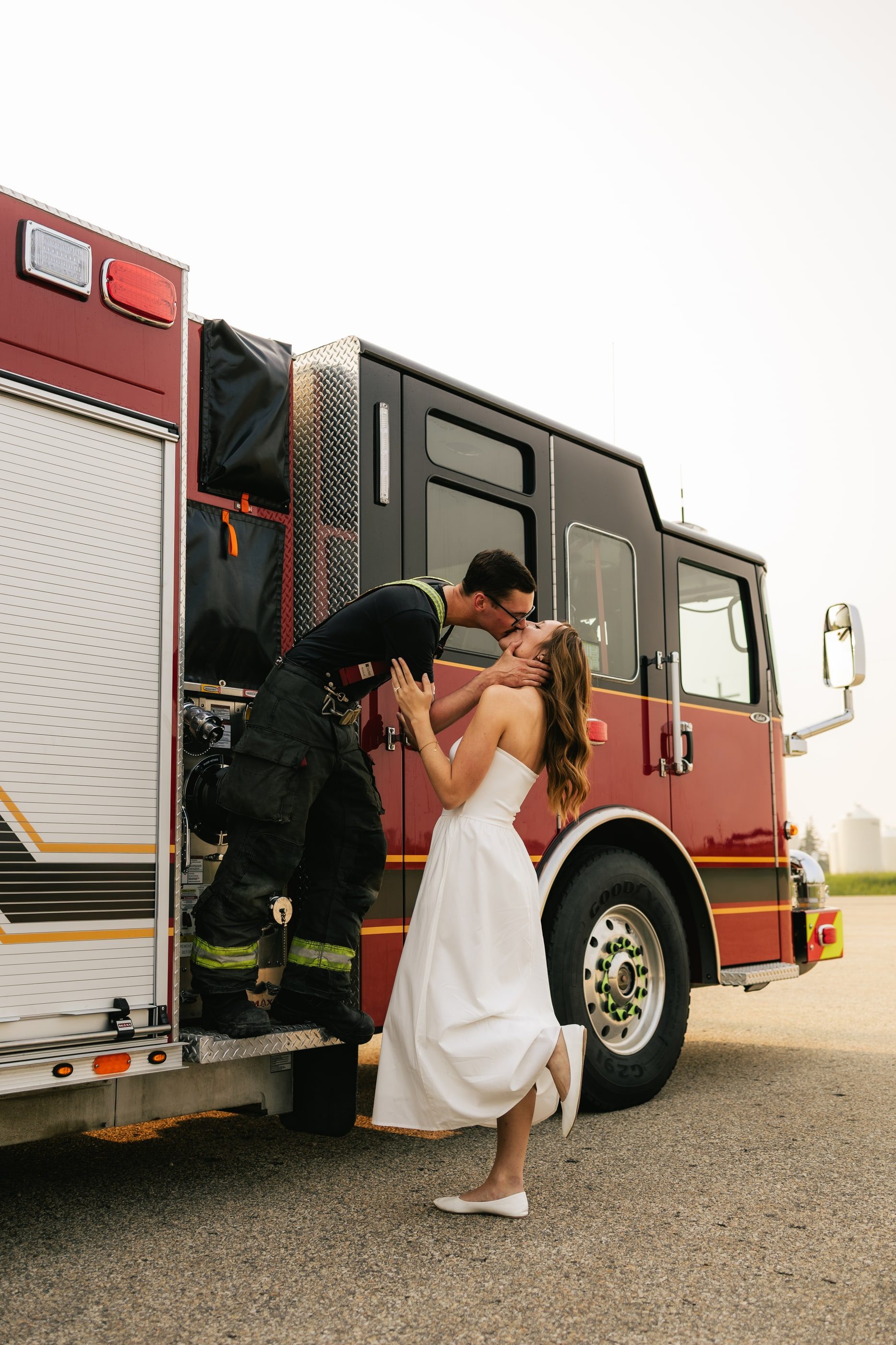 A firefighter leans down from a red fire engine to kiss his partner in a white dress.
