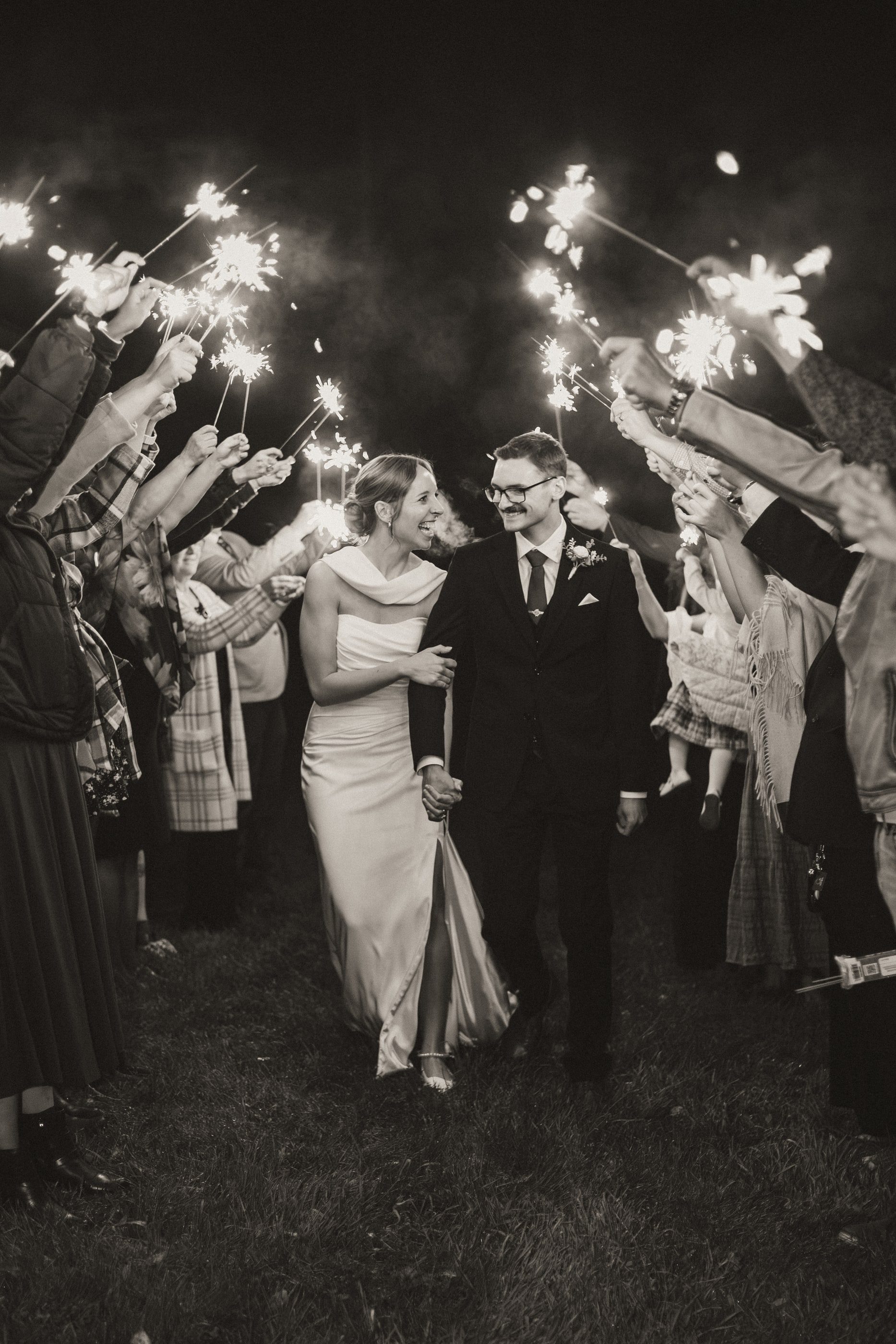 Black-and-white photo of a bride and groom walking through a tunnel of guests holding sparklers.