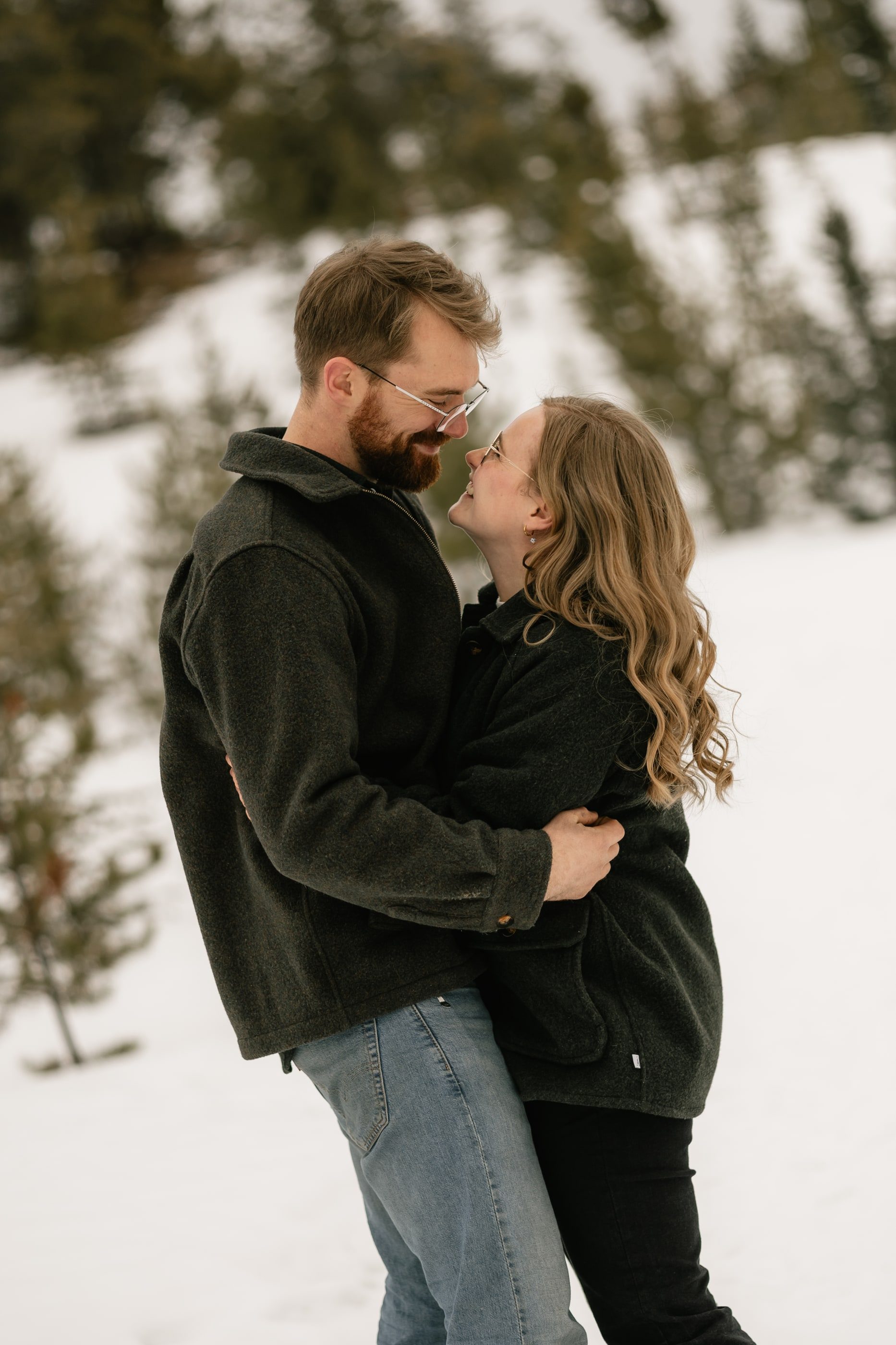 A couple in warm jackets hold each other close and touch noses in a snowy forest.