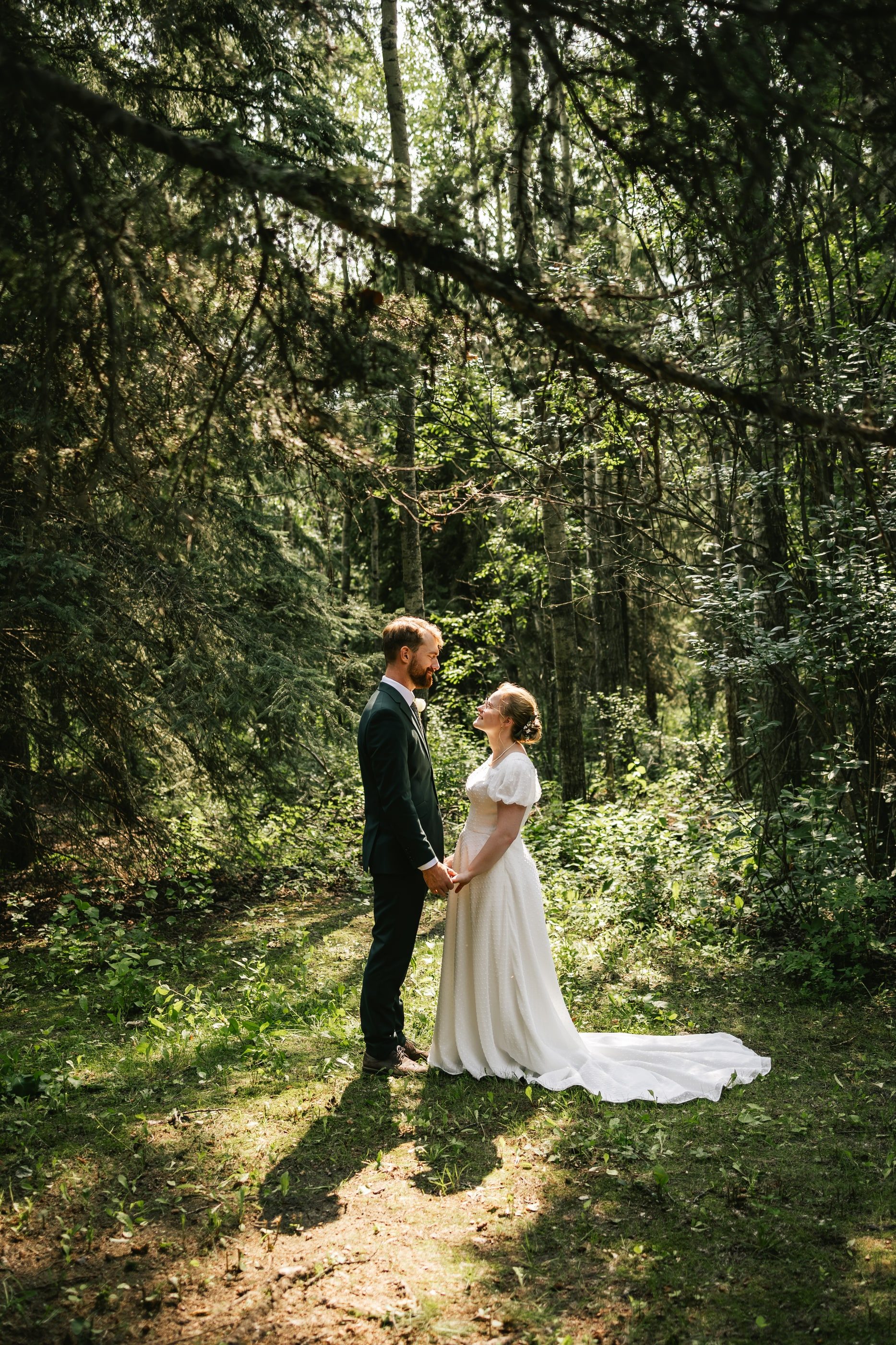A bride and groom hold hands, facing each other in a sun-dappled forest clearing.