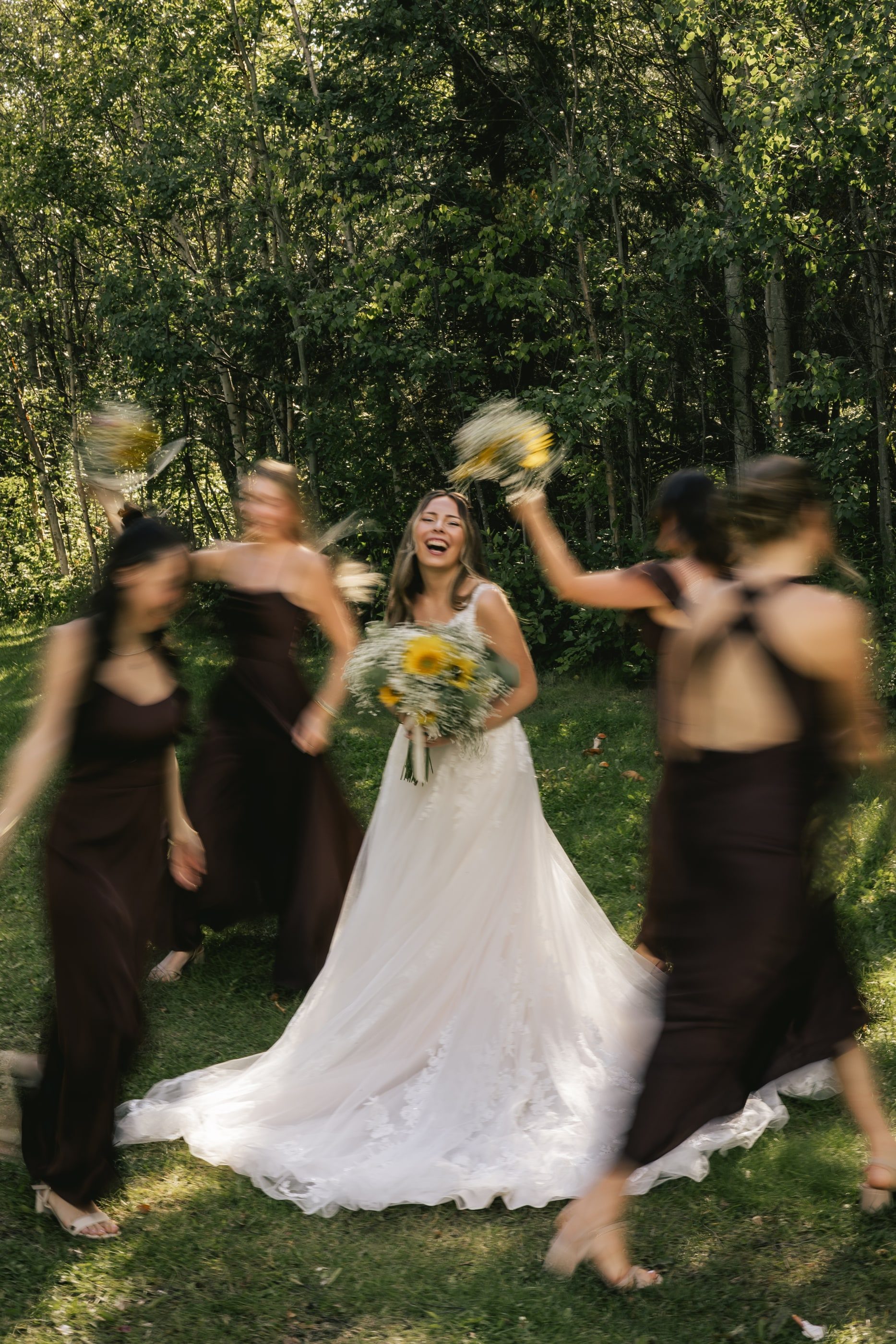 A laughing bride holds her sunflower bouquet as bridesmaids in dark dresses twirl around her.