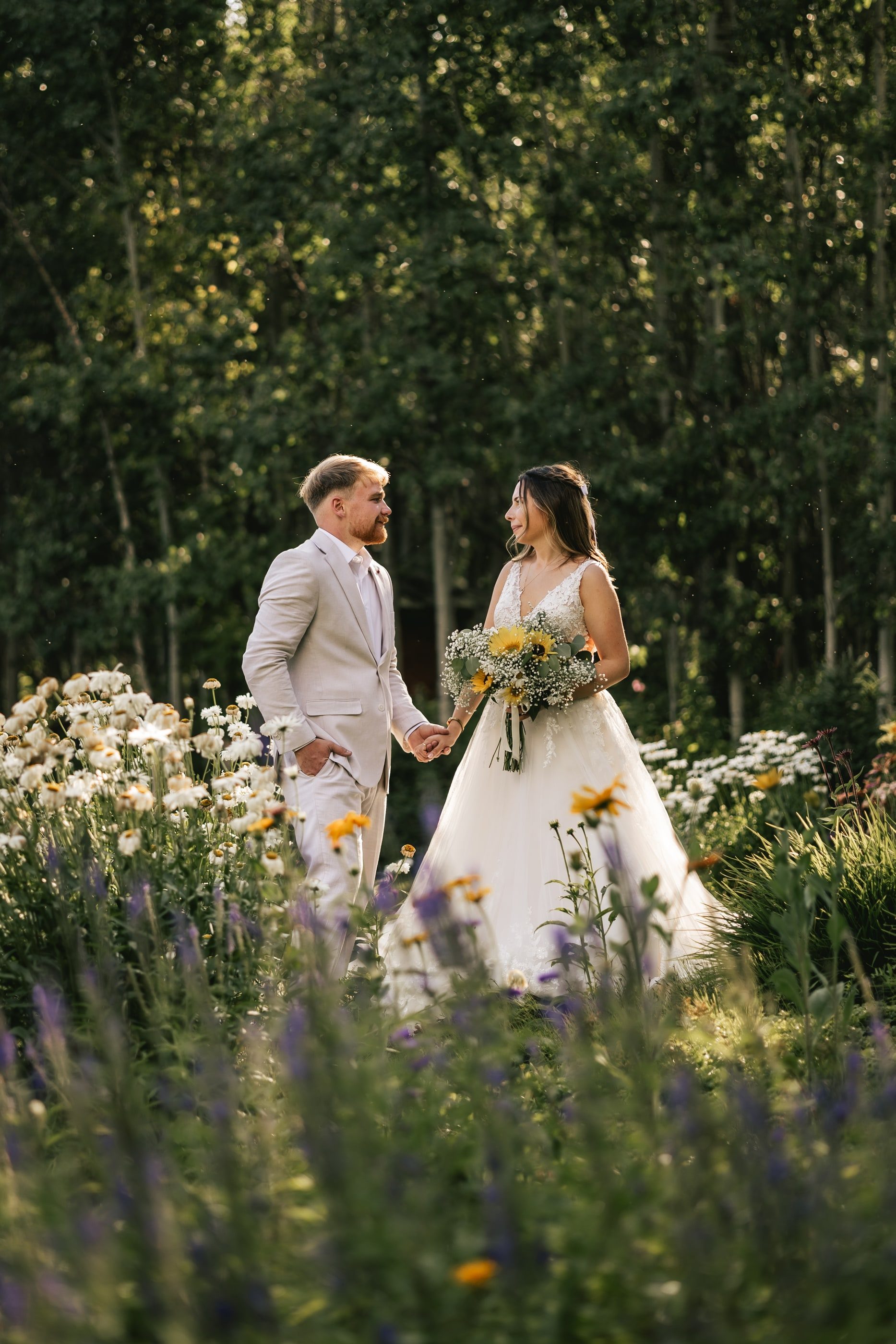 A bride and groom walk through a garden of white daisies and purple wildflowers.