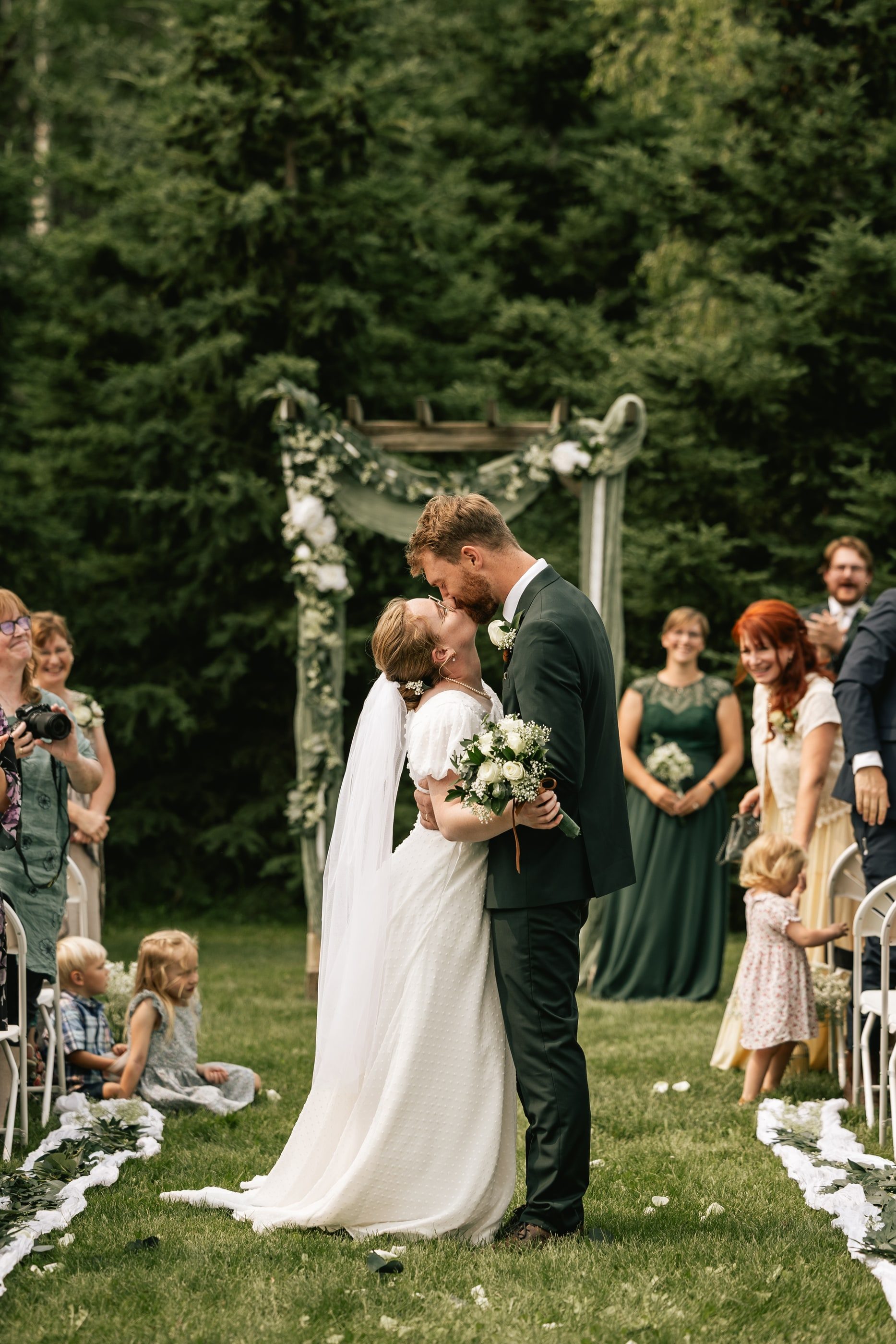 A bride and groom kiss under a floral arch at an outdoor forest ceremony as guests look on.
