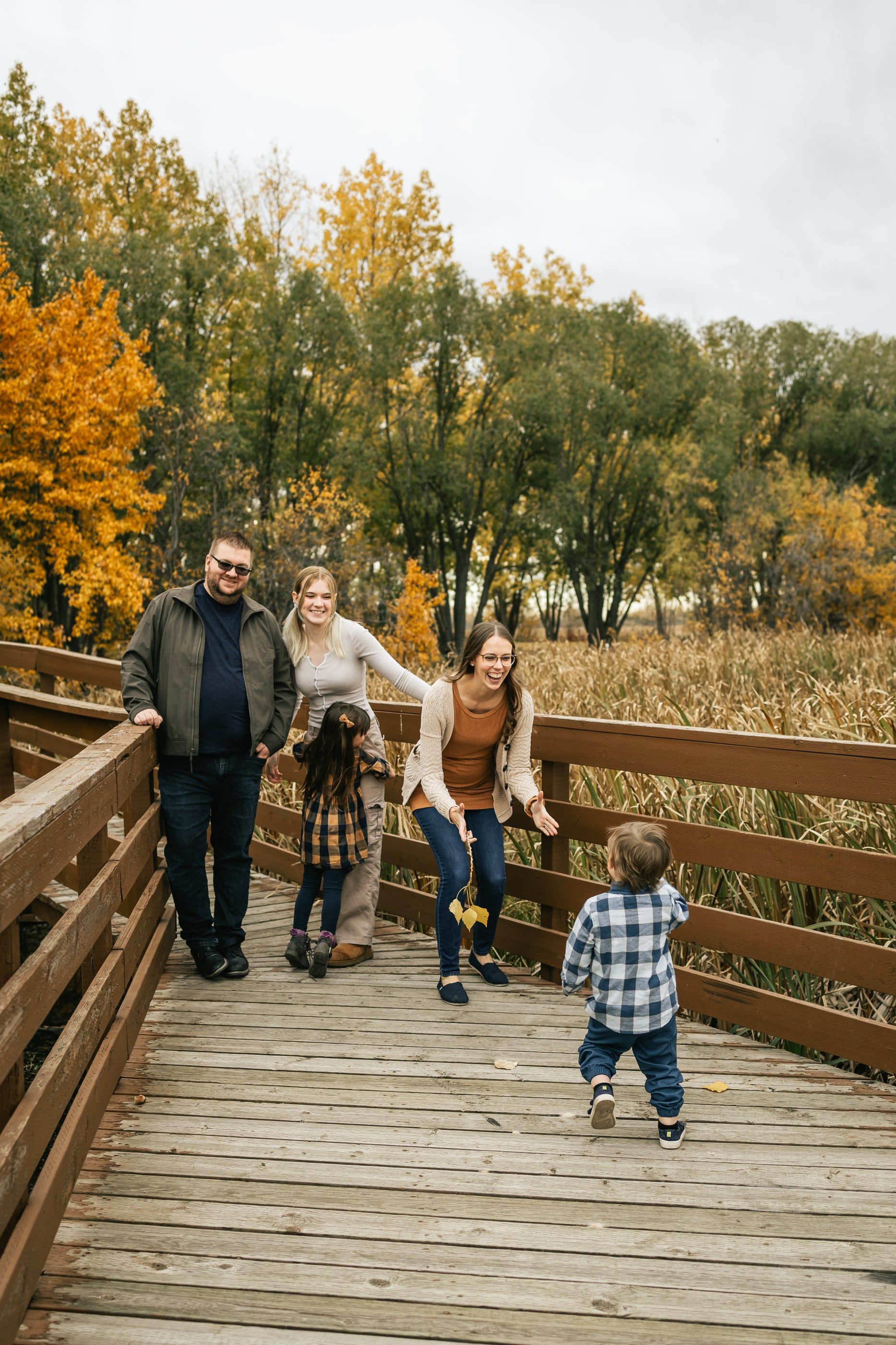 A family walks along a wooden boardwalk through autumn trees as a toddler runs ahead.