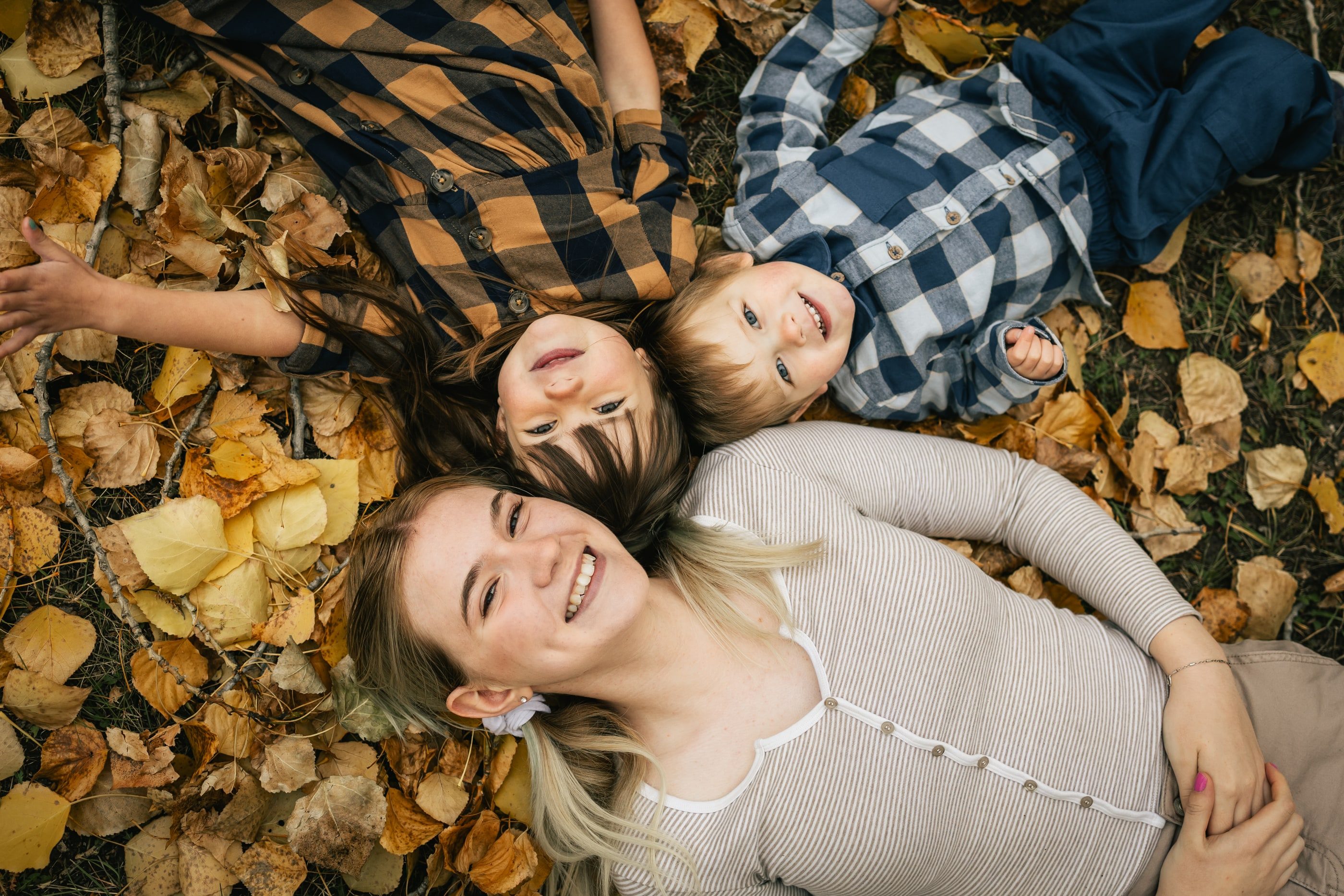 A mother and her two young sons lie head to head on golden autumn leaves, smiling.