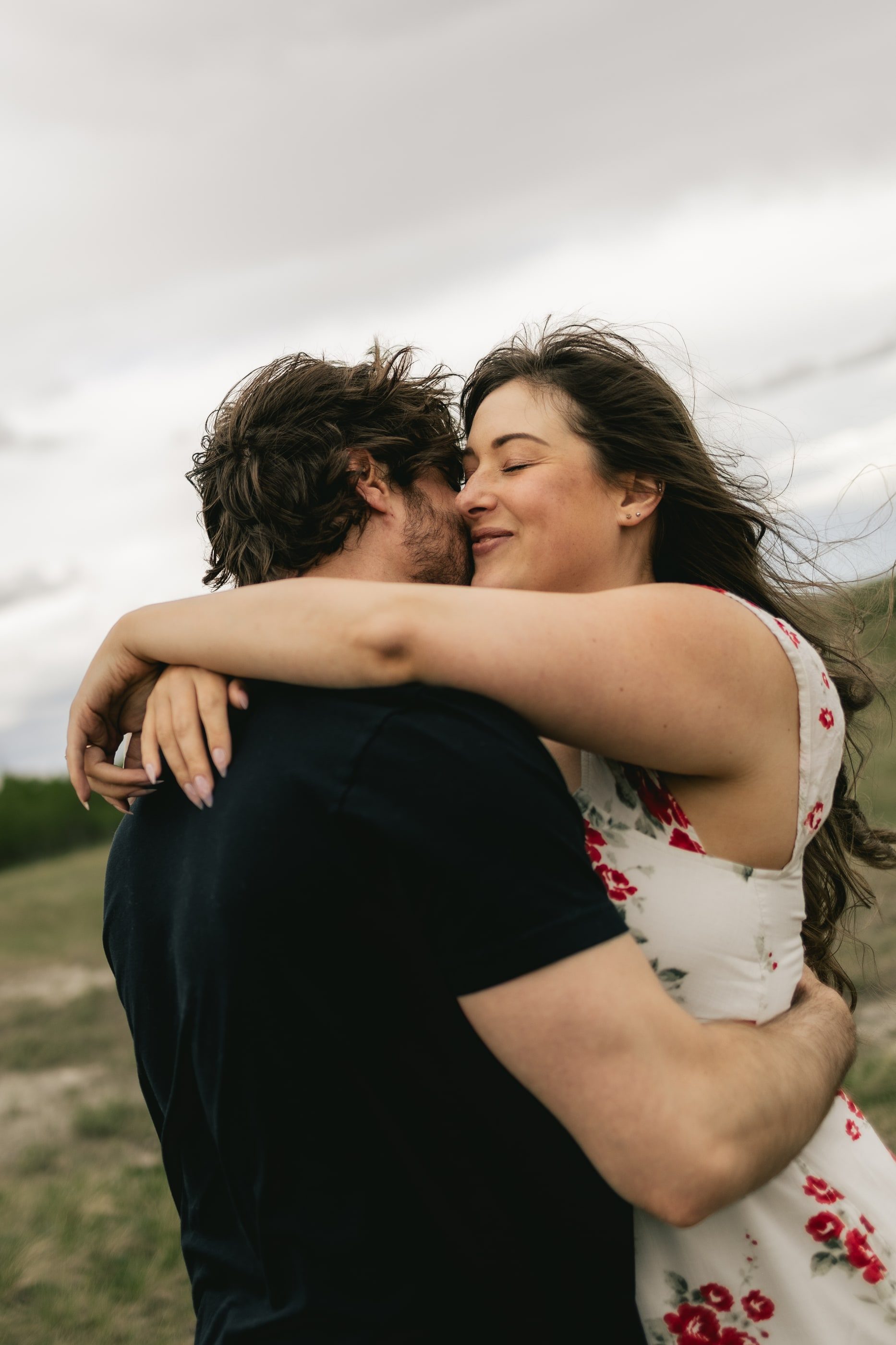 A laughing woman in a red floral dress wraps her arms around her partner's neck under a cloudy sky.
