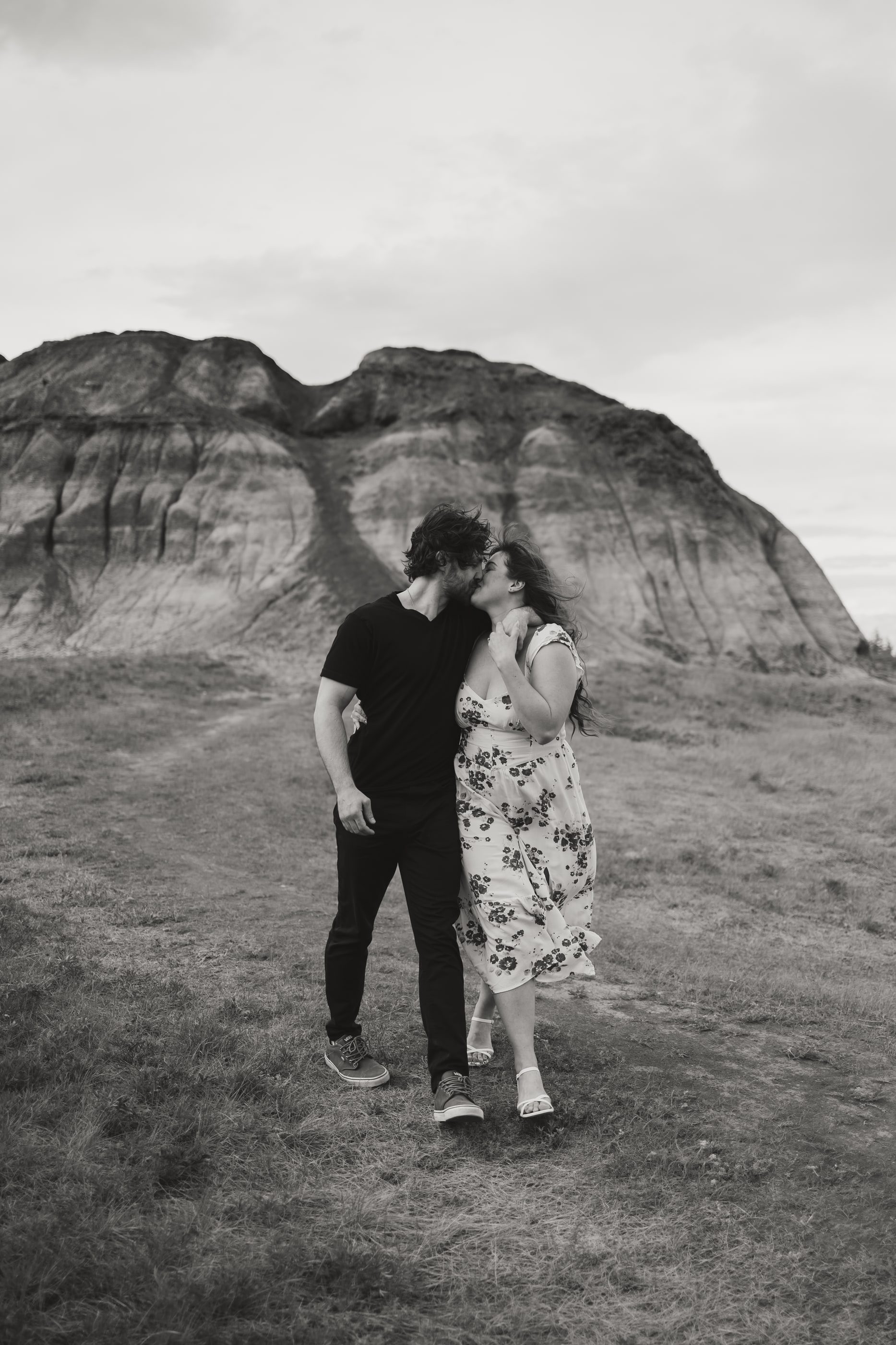 Black-and-white photo of a couple walking hand in hand below a rounded badlands hill.