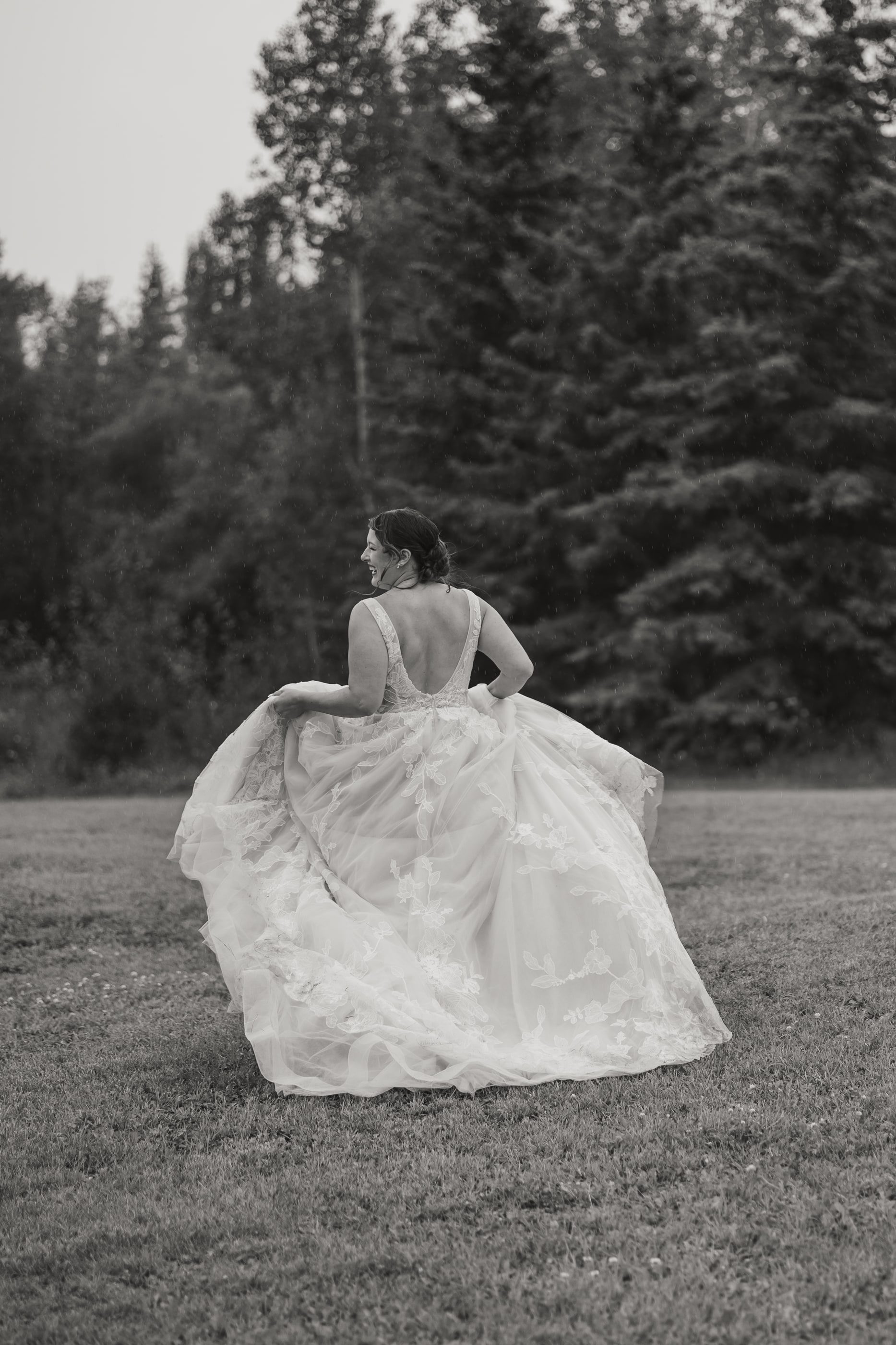 Black-and-white photo of a bride lifting her full skirt as she runs across the grass toward the trees.