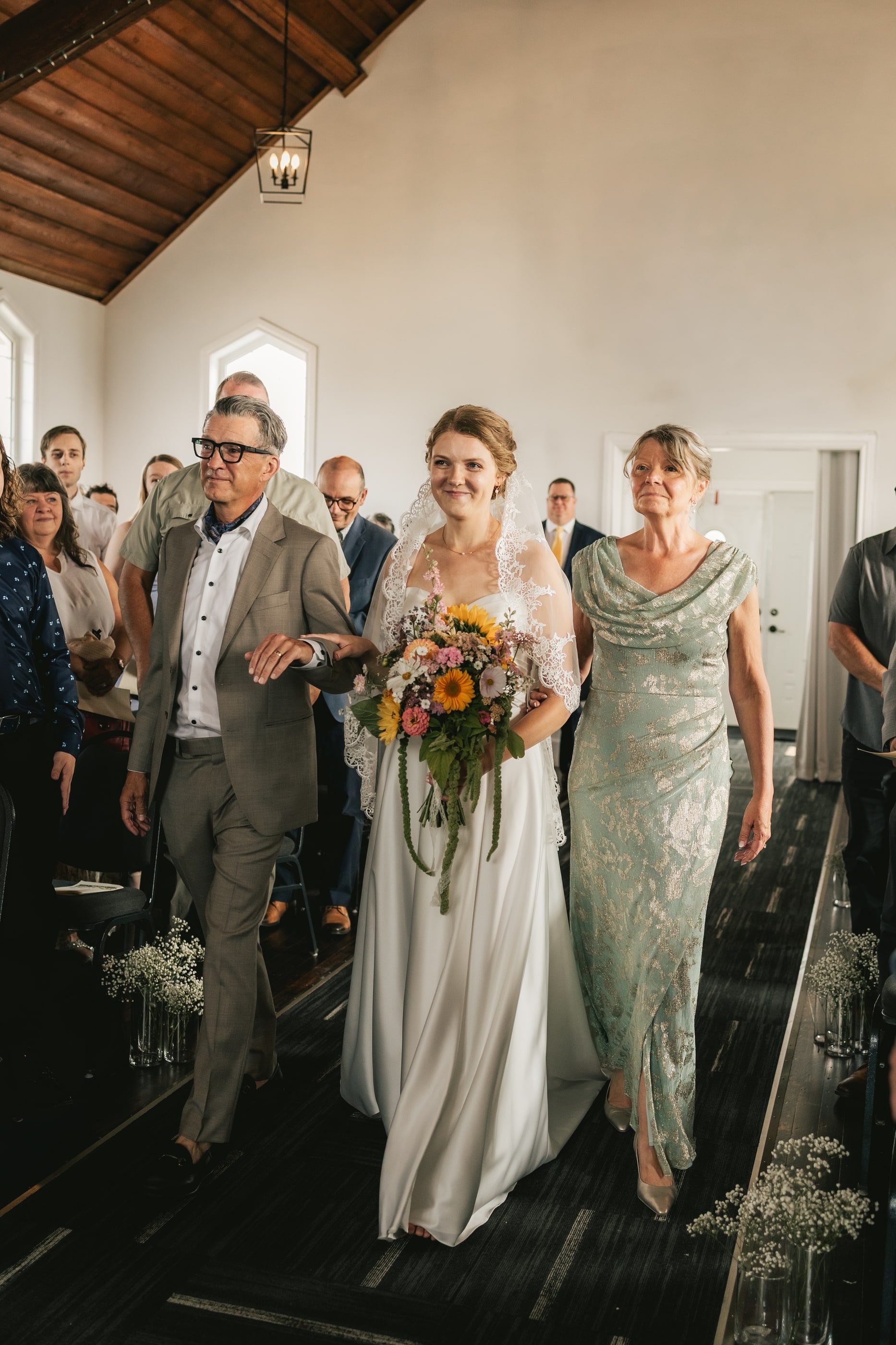A bride walks down the aisle of a white chapel between her parents, holding a sunflower bouquet.