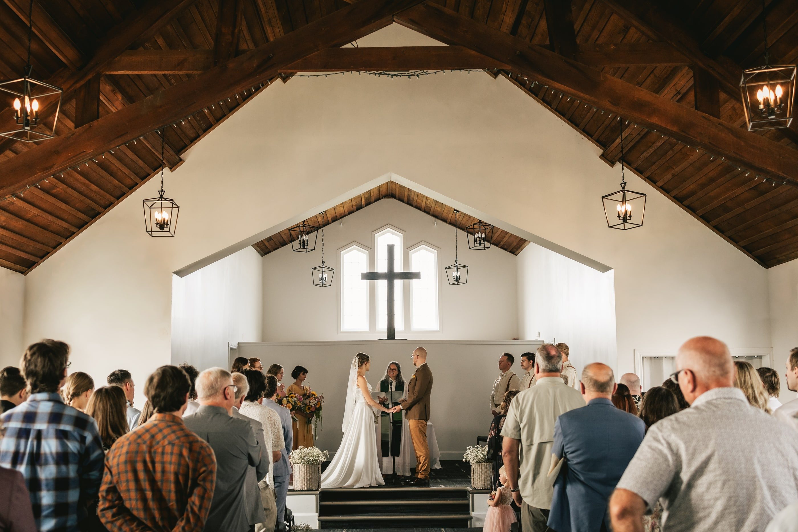 A wedding ceremony inside a white chapel with a vaulted wooden ceiling and a cross above the altar.